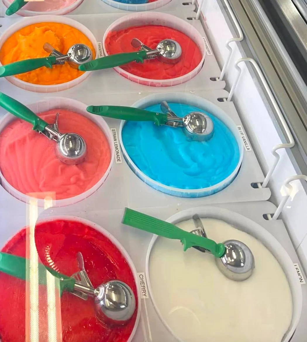 Multiple tubs of colorful ice creams and sorbets, each with a metal scoop and green handle, labeled with flavors like strawberry, cherry, lemon, and vanilla, in an ice cream shop freezer.