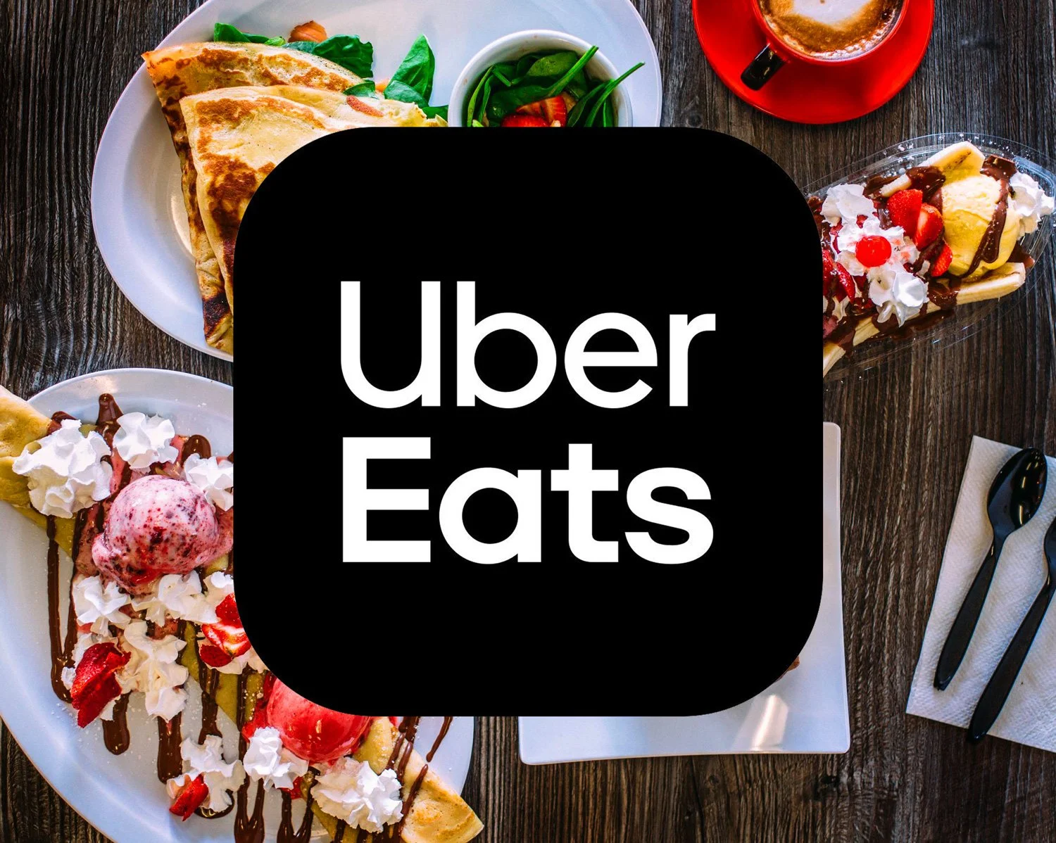 A top-down view of a wooden table with plates of crepes topped with strawberries, chocolate sauce, whipped cream, and ice cream, a cup of coffee in a red mug, and utensils on a napkin, with the Uber Eats logo overlayed