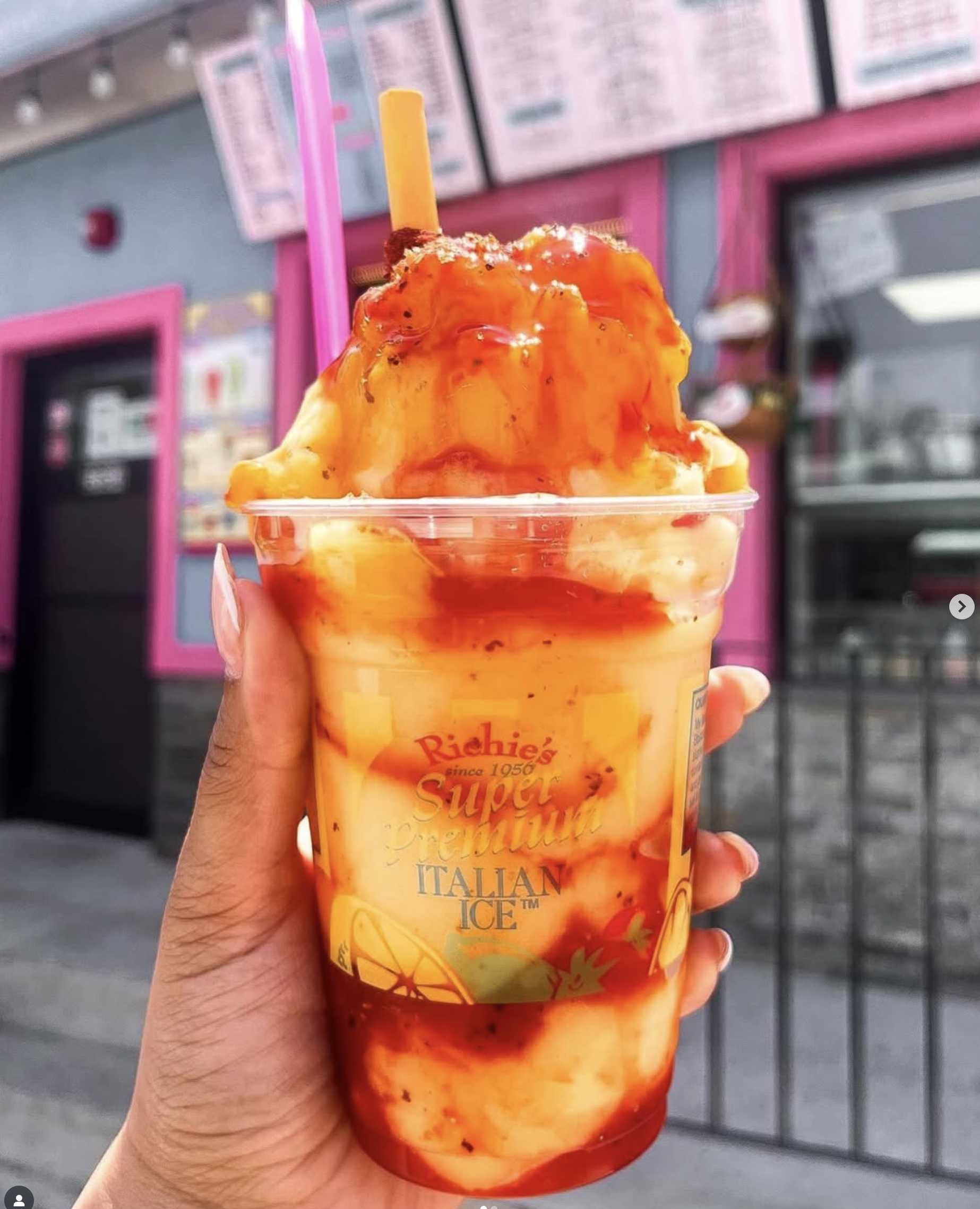 Close-up of a hand holding a cup of mango-flavored Italian ice topped with orange syrup. In the background, there are pink framed menu boards at a shop with pink accents.