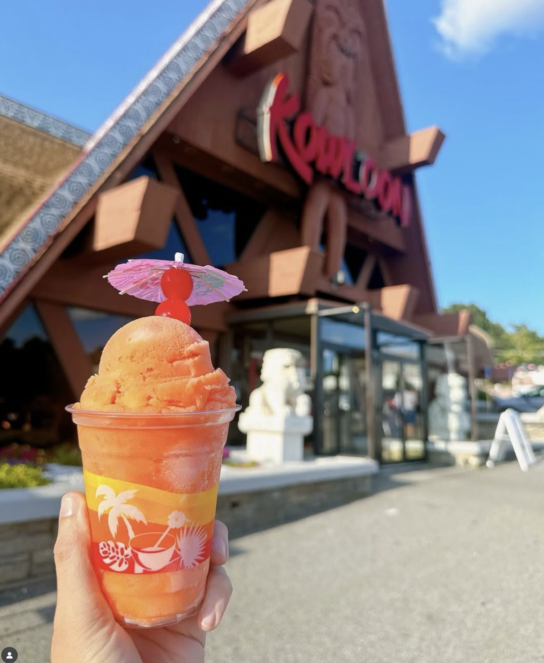 A person holding a colorful tropical-themed ice cream with a pink paper umbrella and red cherries on top, in front of a building with a sign that says 'Kona' on a sunny day.