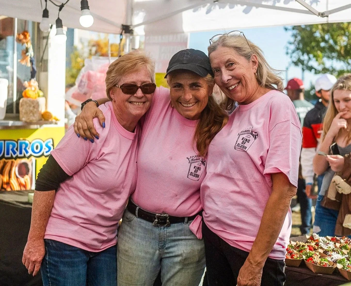 Three women wearing matching pink shirts at an outdoor event, smiling and hugging each other.
