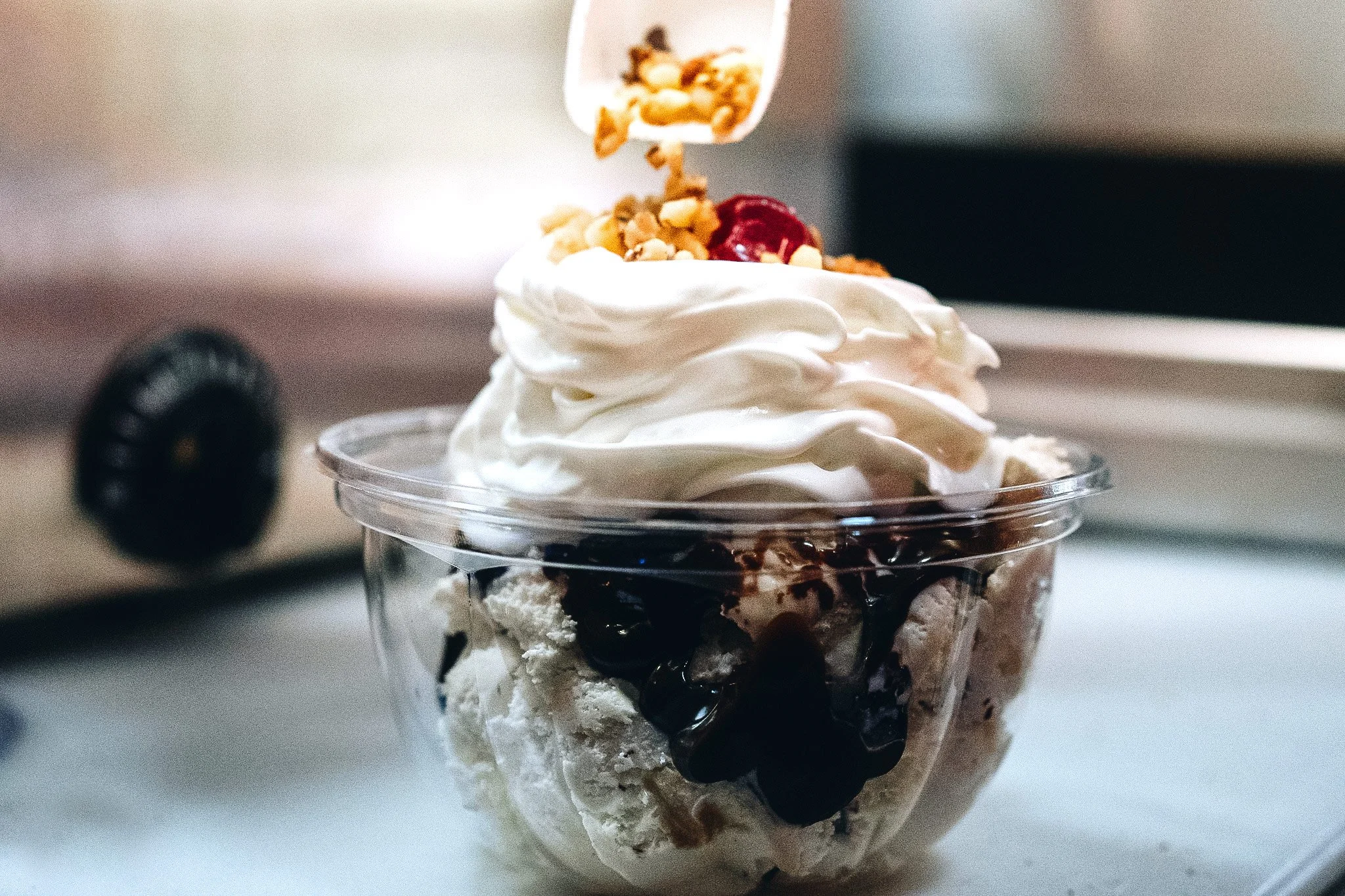 Close-up of a dessert parfait in a clear cup with whipped cream, cherries, syrup, granola, and other toppings.