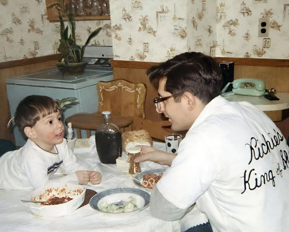 A young boy and a man are sitting at a table having a meal. The boy looks at the man with interest, and the man is smiling and holding a sandwich. The table has food, including spaghetti, salad, and a bottle of dark soda. The man is wearing glasses and a white shirt with the words 'Richie's King of' written on the back.
