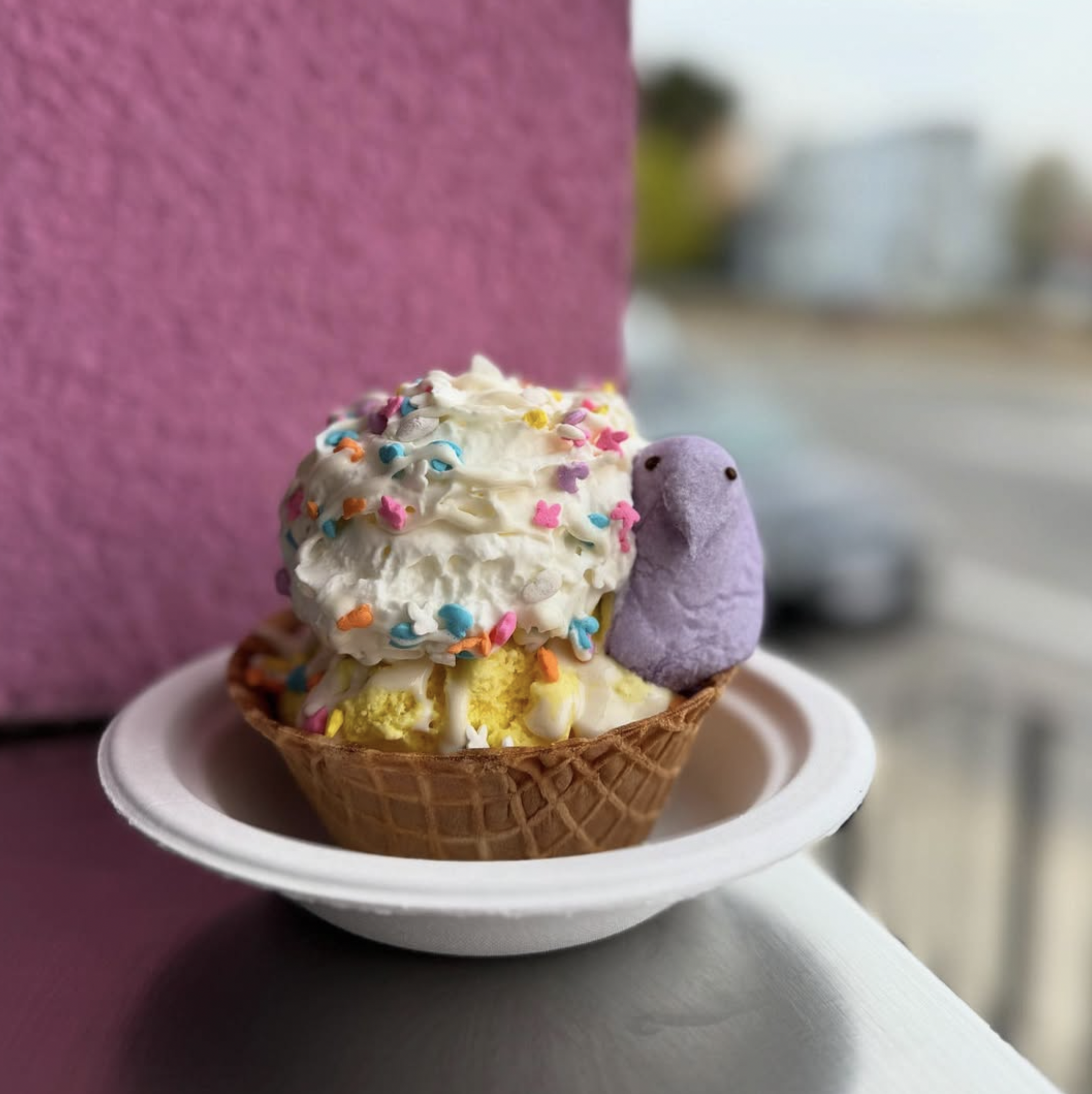 Ice cream cone with white ice cream, colorful sprinkles, and a purple bear-shaped marshmallow, placed on a white plate.