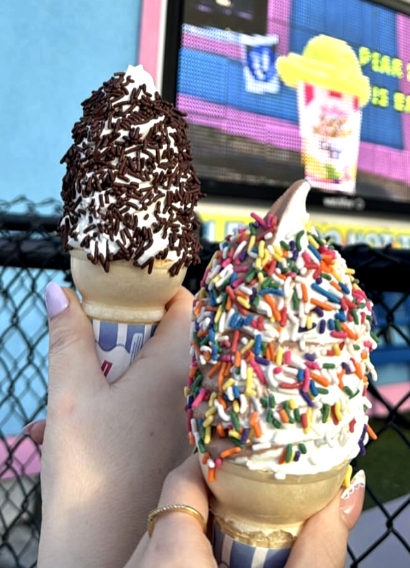 Two ice cream cones held up in front of a colorful background, one with chocolate sprinkles and the other with rainbow sprinkles.