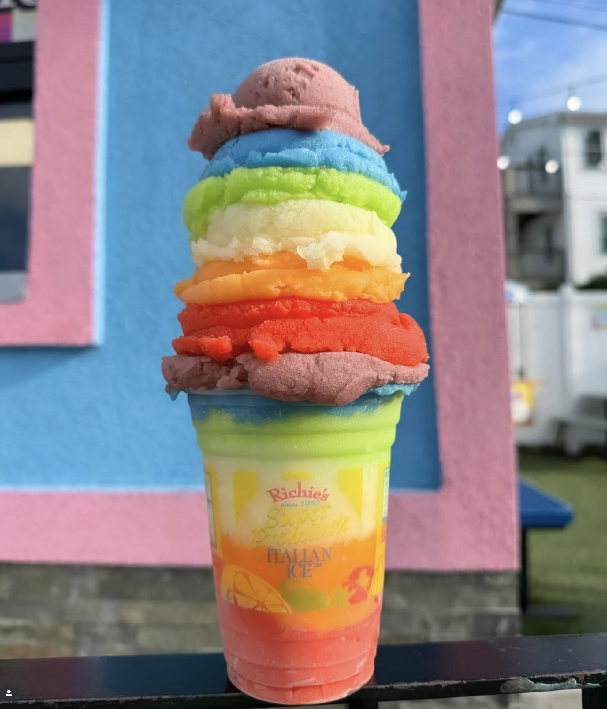 A rainbow-colored cone with six scoops of ice cream stacked vertically. The scoops are pink, blue, green, white, orange, and red from top to bottom. The cone has the logo "Richie's" and the words "since 1950" on it. The background includes pink and b