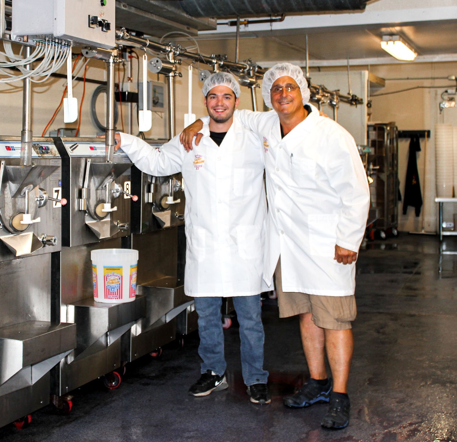 Two men in white coats and hairnets standing next to large ice cream machines inside an ice cream production facility, smiling for the camera.