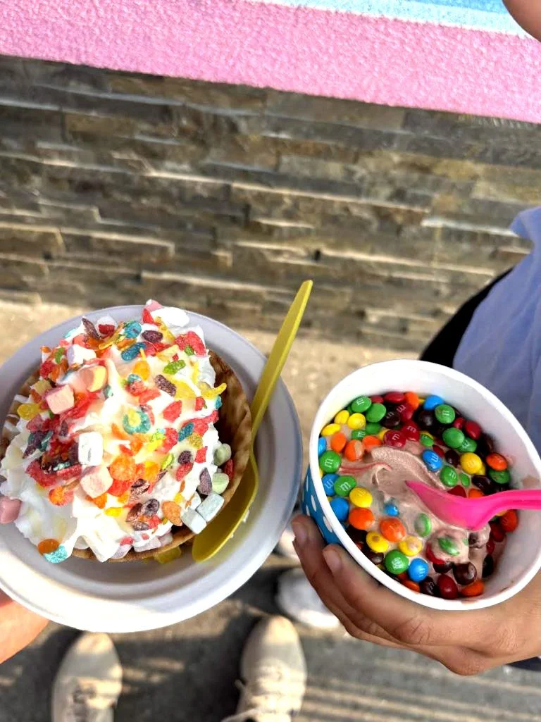 Two cups of ice cream topped with colorful candies and sprinkles, held outdoors near a stone wall.