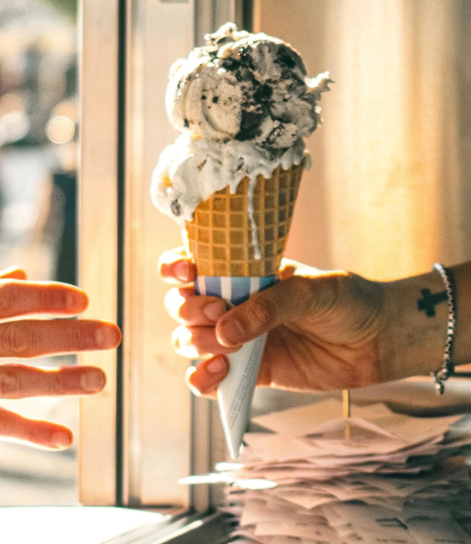 Close-up of a hand holding an ice cream cone with two scoops of vanilla ice cream topped with chocolate syrup, in front of a window with sunlight streaming in.
