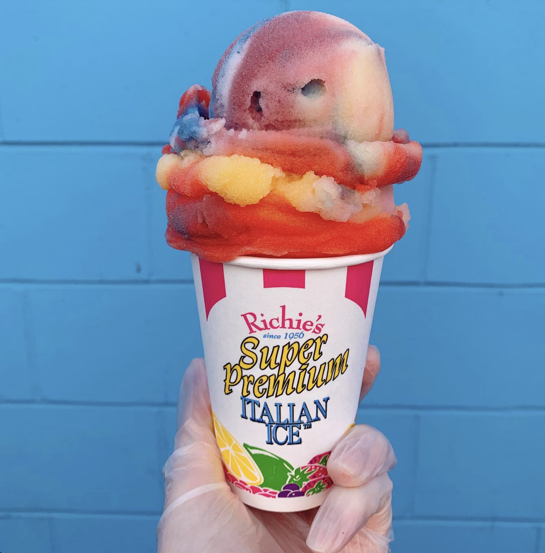 Close-up of a person's hand holding a cup of colorful ice cream against a blue background. The cup is branded with 'Richie's Summer Super Premium Italian Ice' and features illustrations of fruit.
