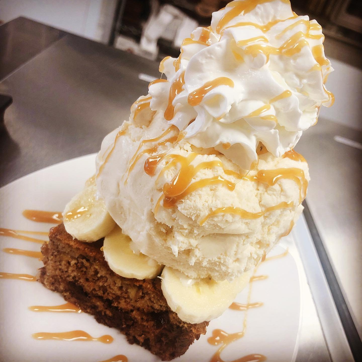 A dessert featuring a slice of chocolate cake topped with banana slices, a scoop of vanilla ice cream, whipped cream, and caramel drizzle.