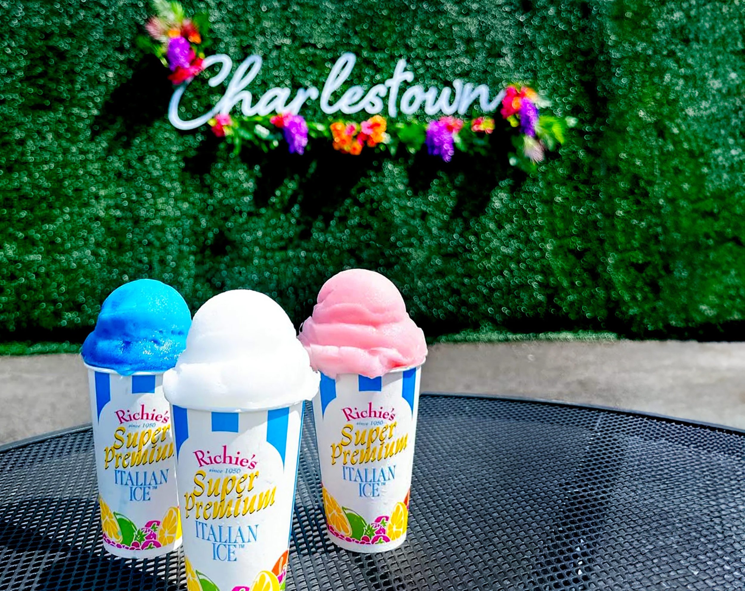 Four cups of Italian Ice with colorful flavors from Richie’s Super Premium, placed on a black metal table outdoors with green hedges and a neon Charleston sign in the background.