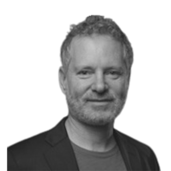 Black and white portrait of a man with curly hair and a beard, wearing a blazer and a T-shirt.