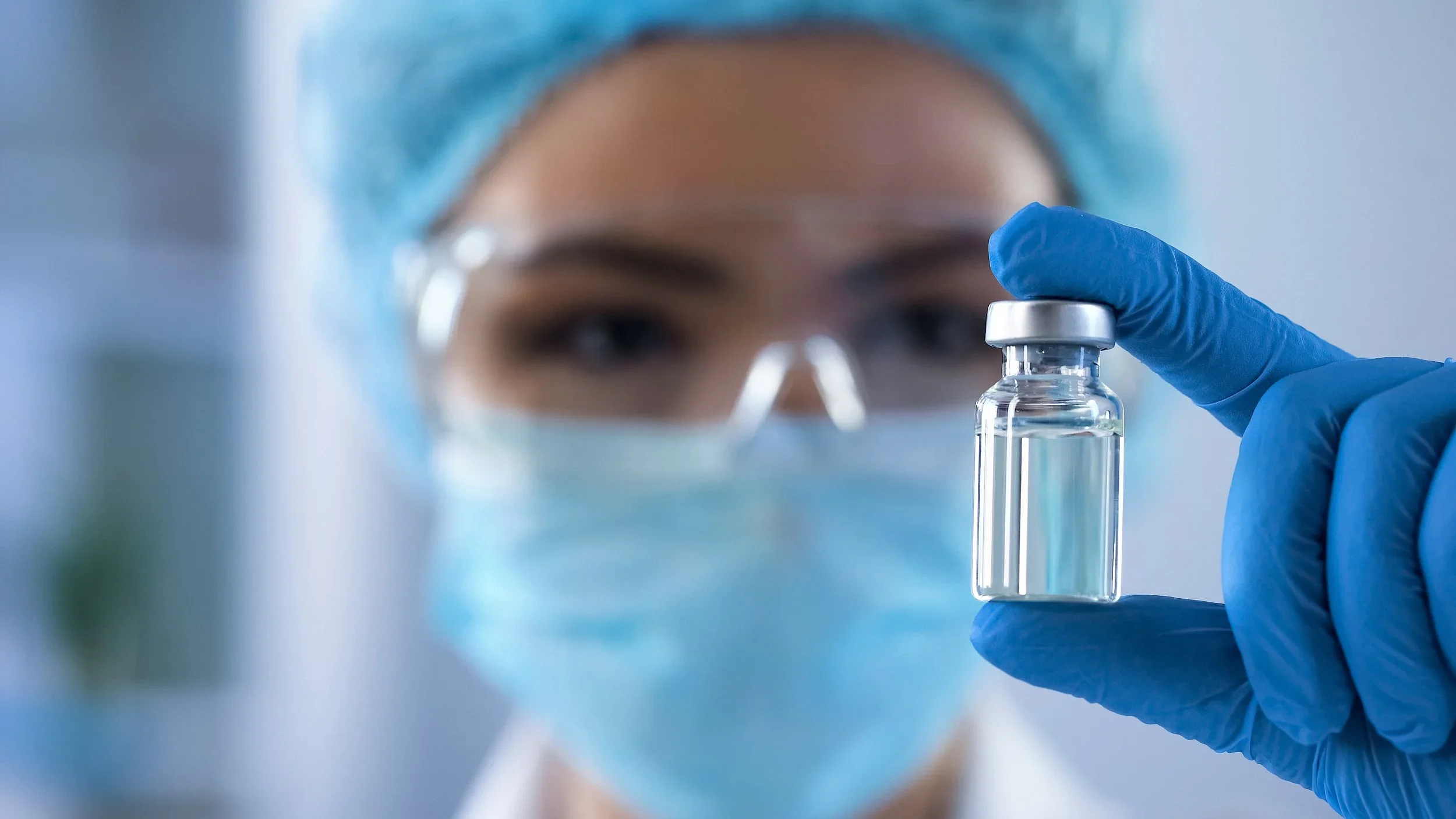 Close-up of a scientist wearing protective gear, including a face mask, goggles, and a hair cover, holding a small glass vial filled with a clear liquid.