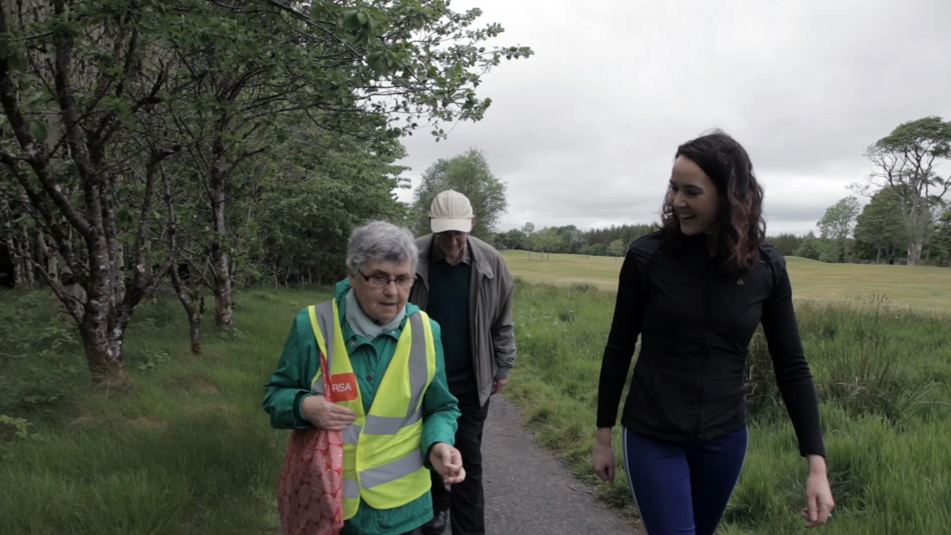 Three people walking outdoors on a grassy trail, with trees on the left and an open field on the right. They are dressed in casual and sportswear, with one person wearing a reflective vest and hat, another in a jacket and hat, and a woman in a black athletic jacket. The weather appears overcast.