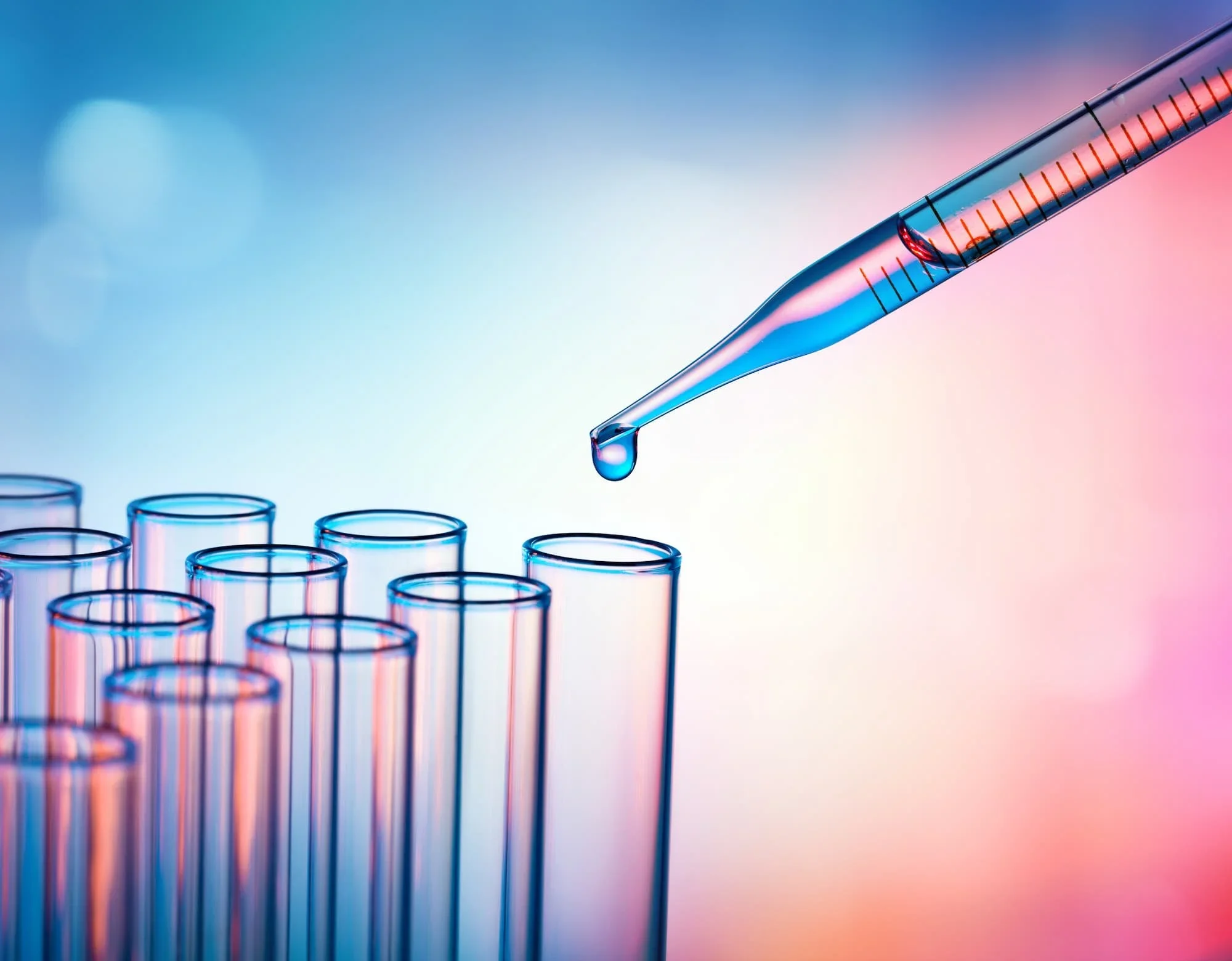 Close-up of a glass micropipette with a droplet of liquid hanging from its tip, over a row of graduated test tubes with colorful reflections and a gradient background.