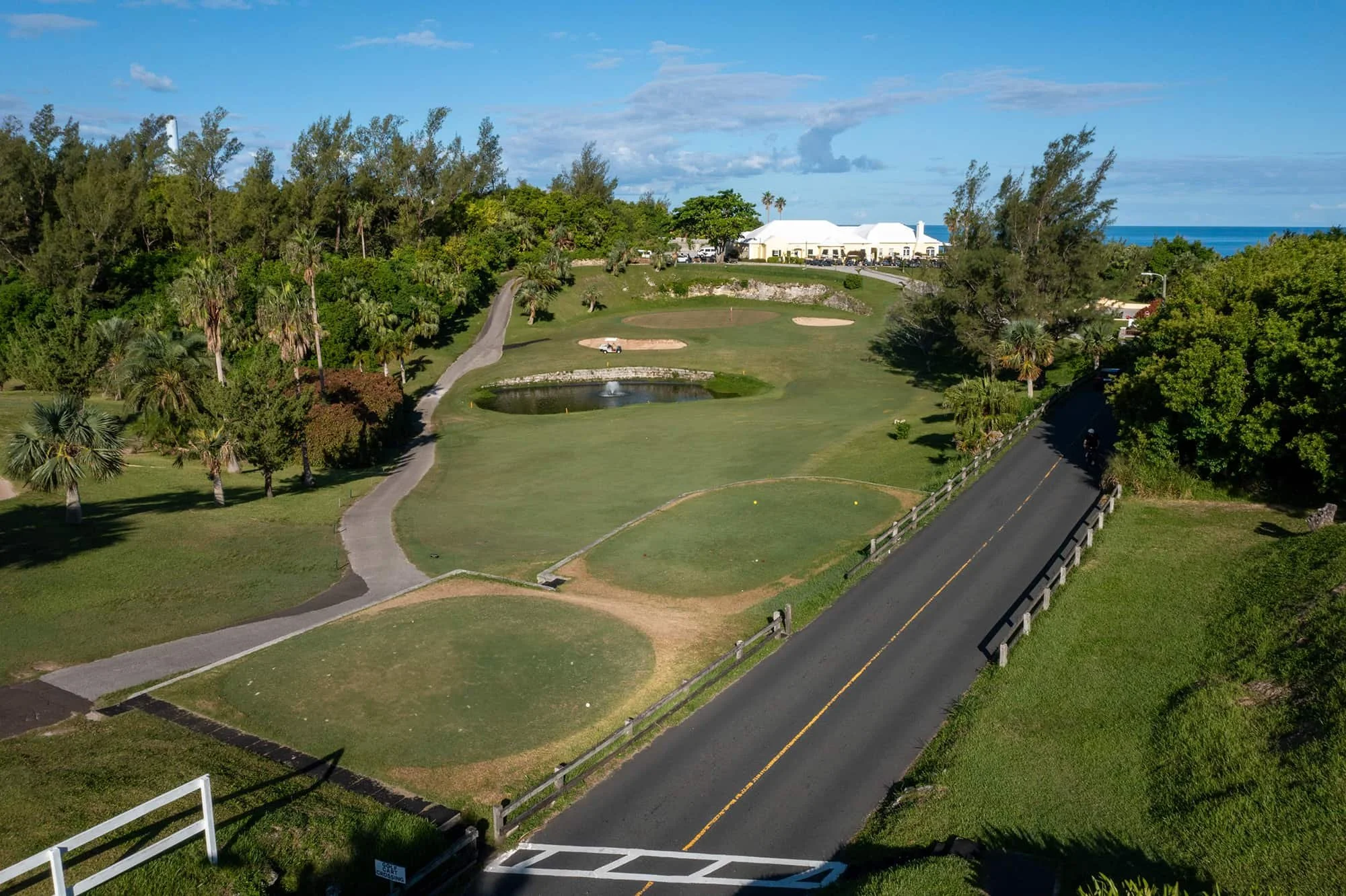 Aerial view of a golf course with a pond, sand traps, and lush green trees, near the coastline with the ocean visible in the background.