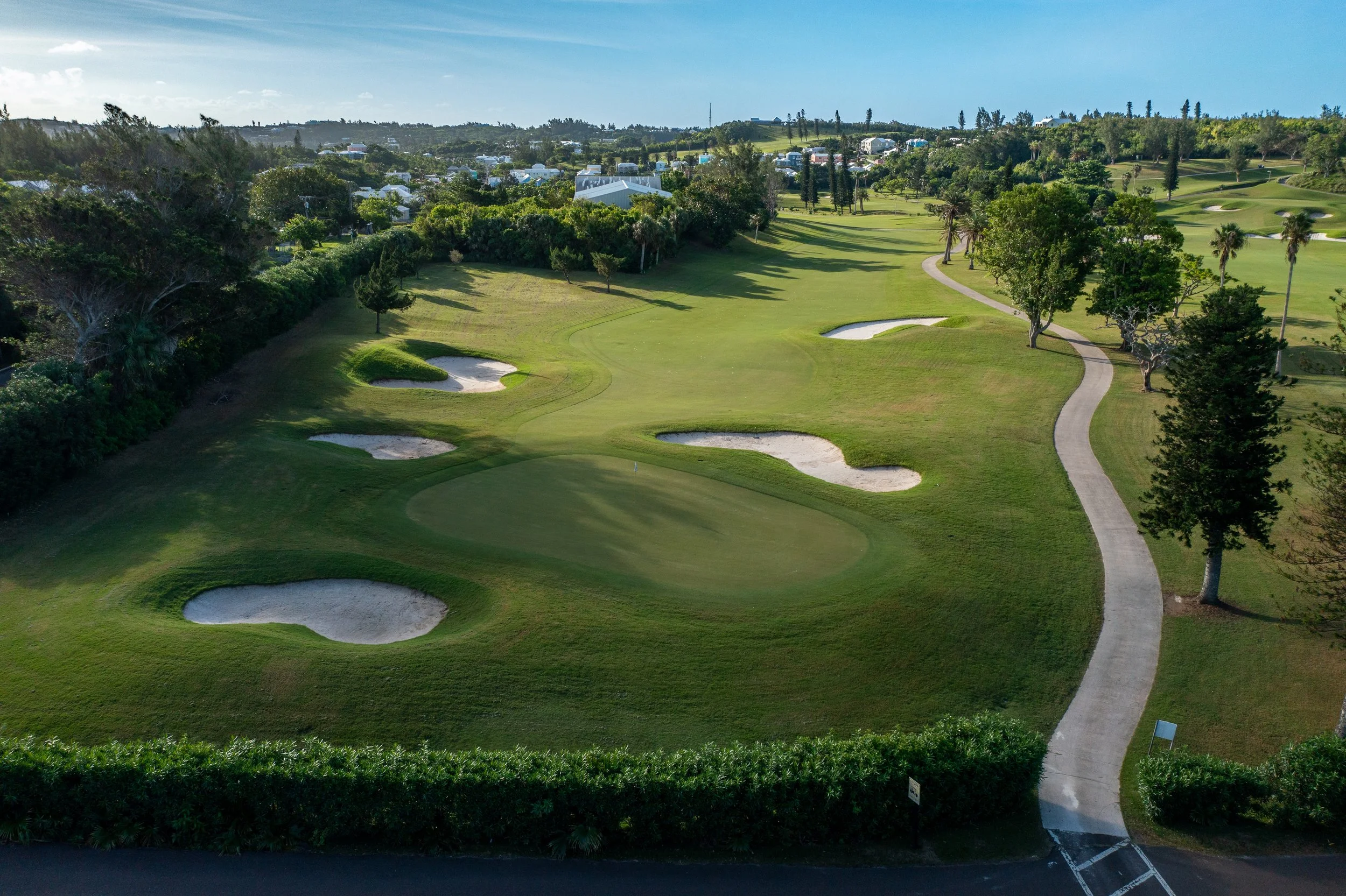 Aerial view of a golf course with sand bunkers, trees, and a walking path, surrounded by residential homes and greenery.
