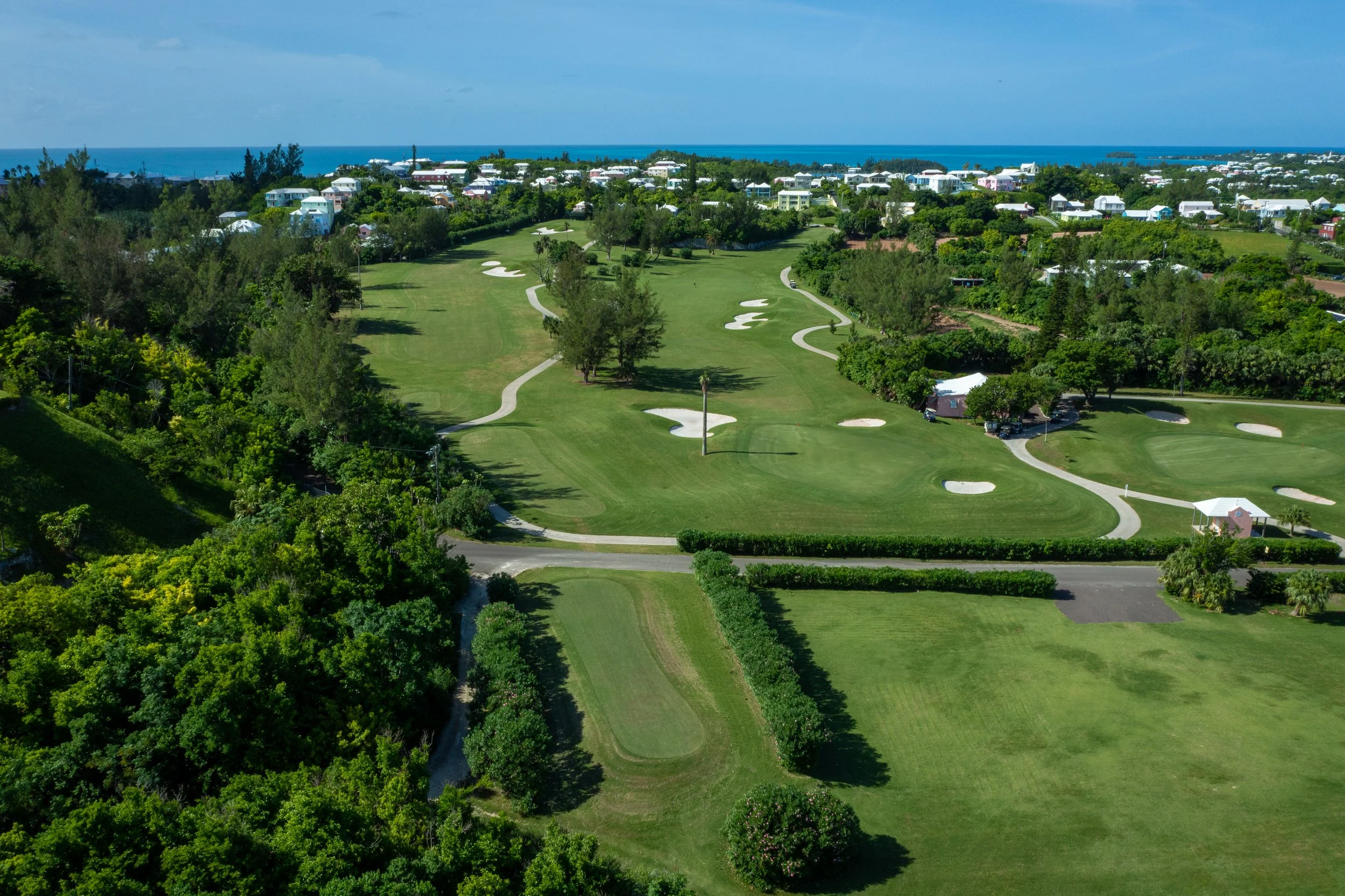 Aerial view of a golf course with green fairways, sand traps, surrounded by trees, with residential houses in the background and the ocean in the distance under a partly cloudy sky.