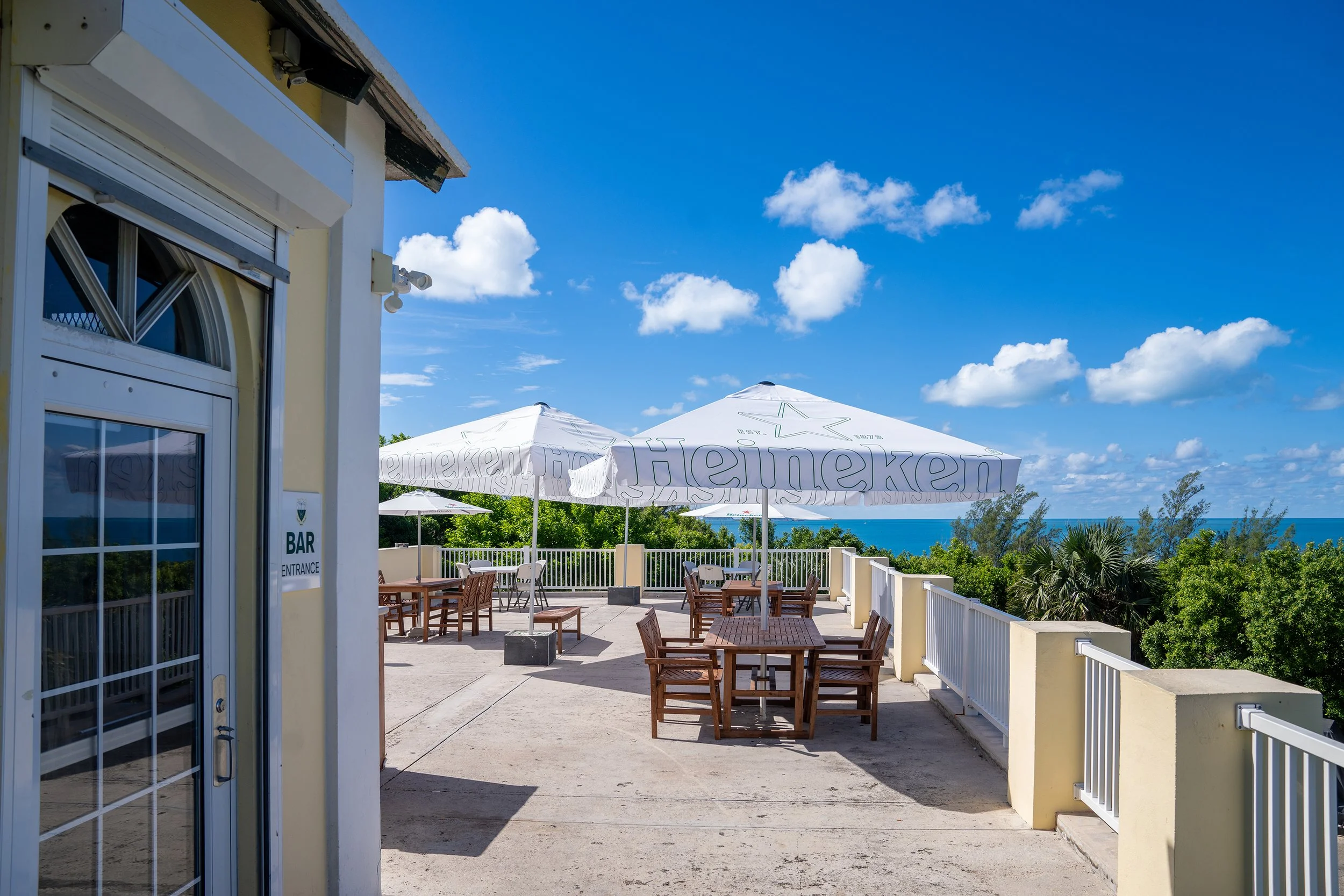 An outdoor patio area with wooden tables and chairs, shaded by Heineken umbrellas, overlooking a lush green landscape and the ocean under a bright blue sky with scattered white clouds.