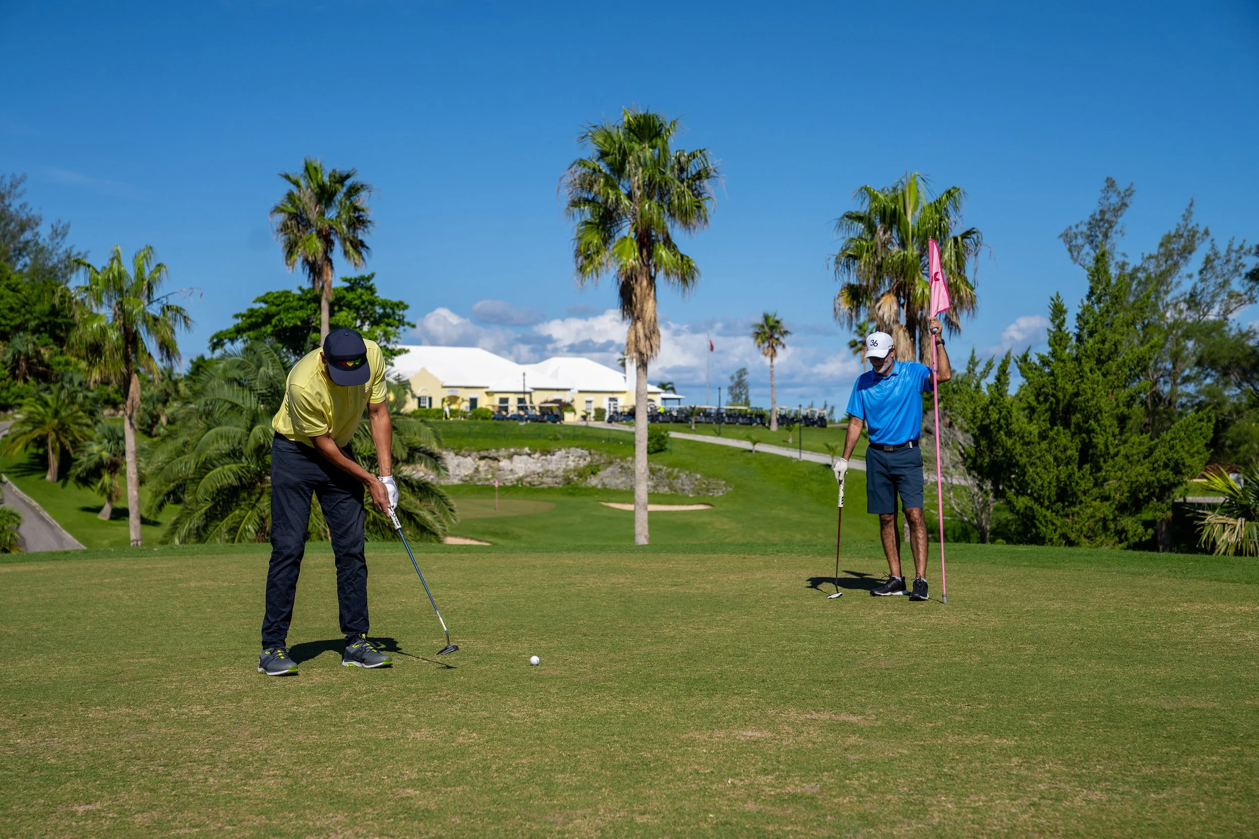 Two men playing golf on a green field with palm trees and a white building in the background on a sunny day.