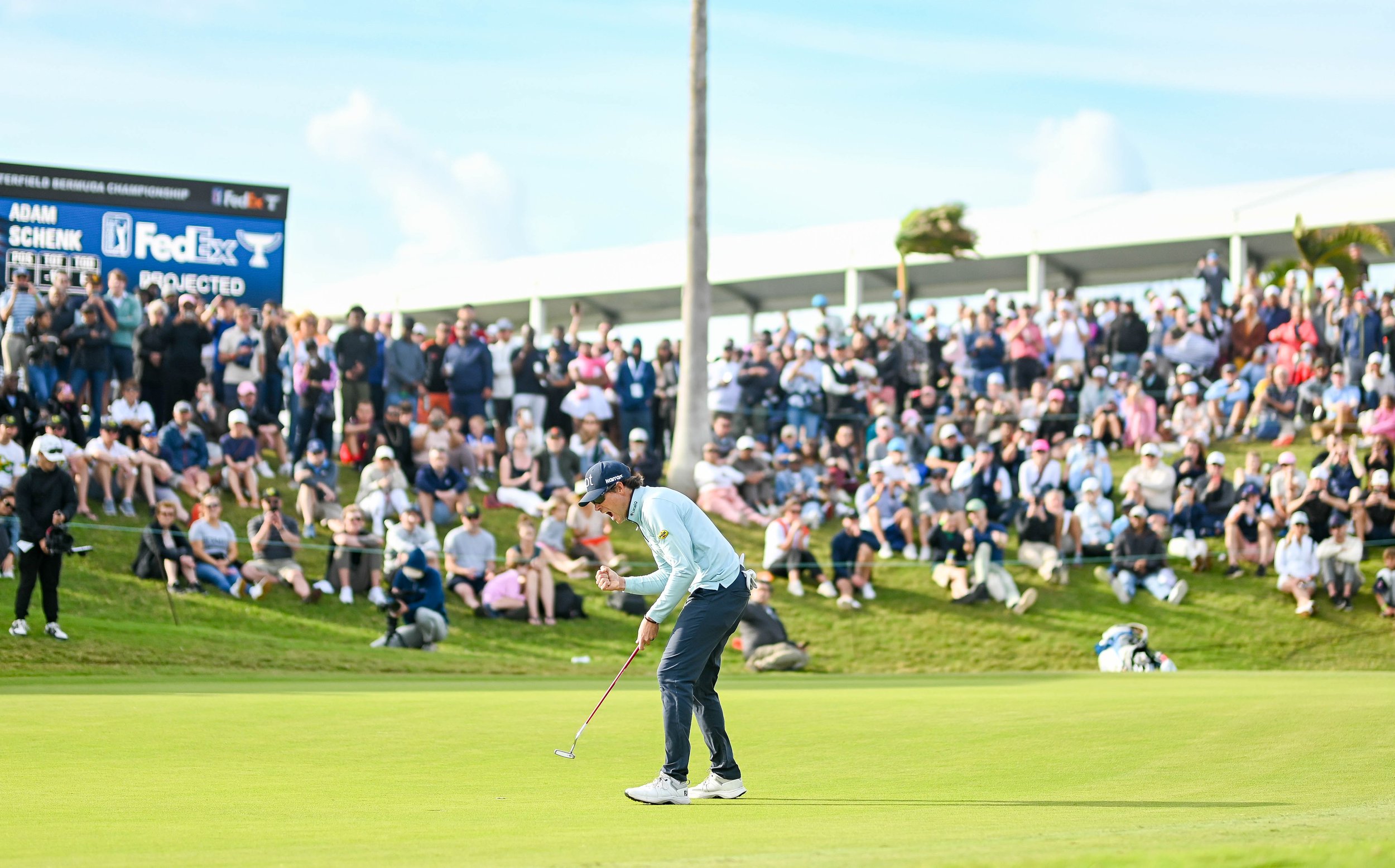A professional golfer putting on a golf course with a large crowd of spectators watching behind him and a scoreboard in the background.