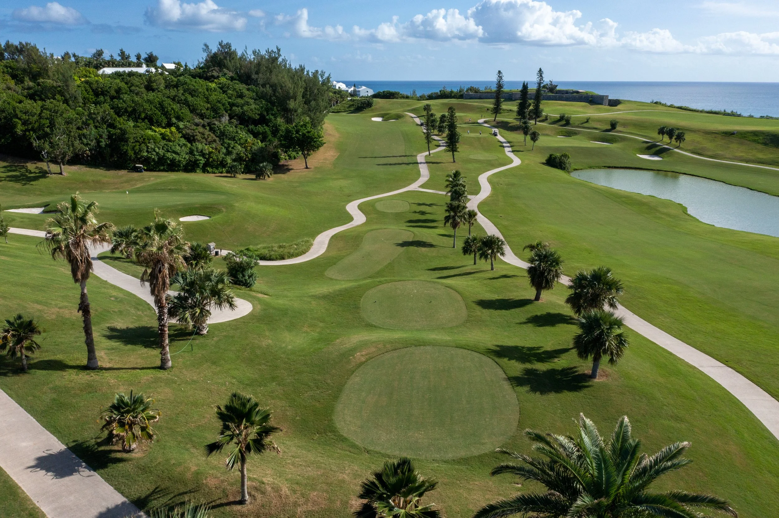 A scenic view of a golf course overlooking the ocean, with a winding path, trees, water hazards, and a blue sky with scattered clouds.