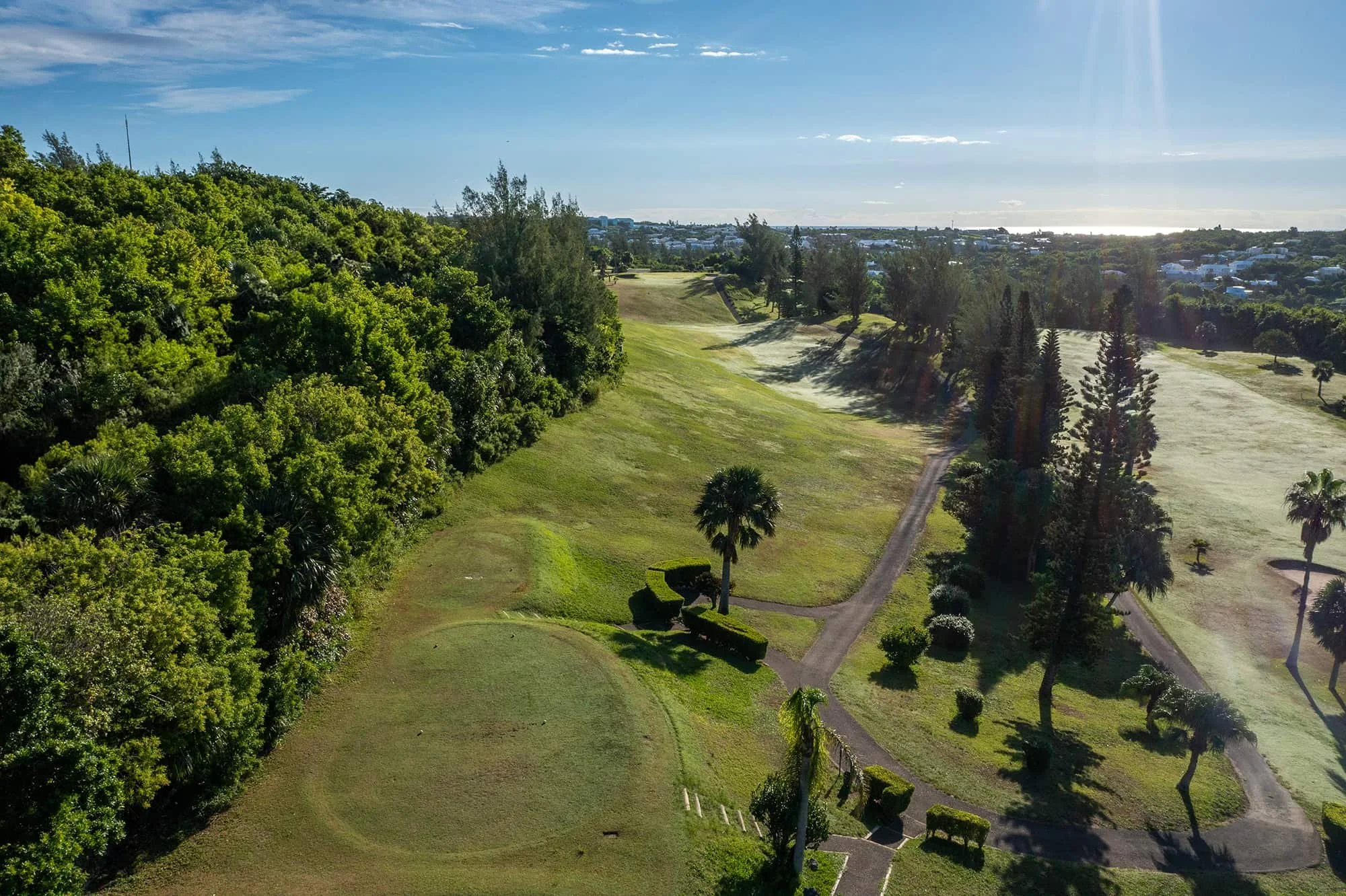 Aerial view of a golf course with green grass, sand bunkers, trees, and a pathway, with a cityscape and water in the distance under a partly cloudy sky.