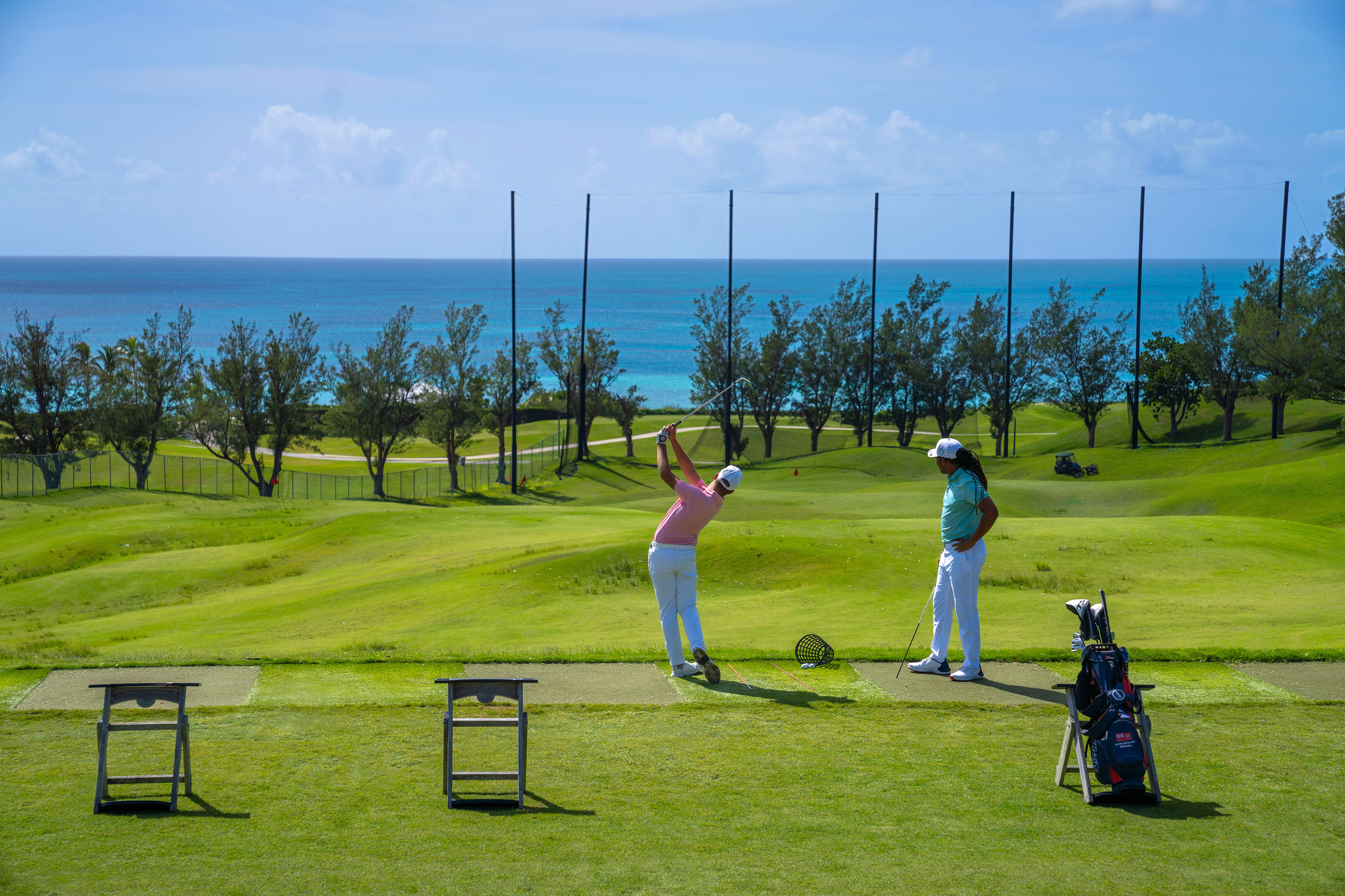 Two women playing golf on a course with lush green grass, trees, and a view of the ocean in the background. One woman is swinging a club while the other watches. Golf equipment and benches are visible in the foreground.