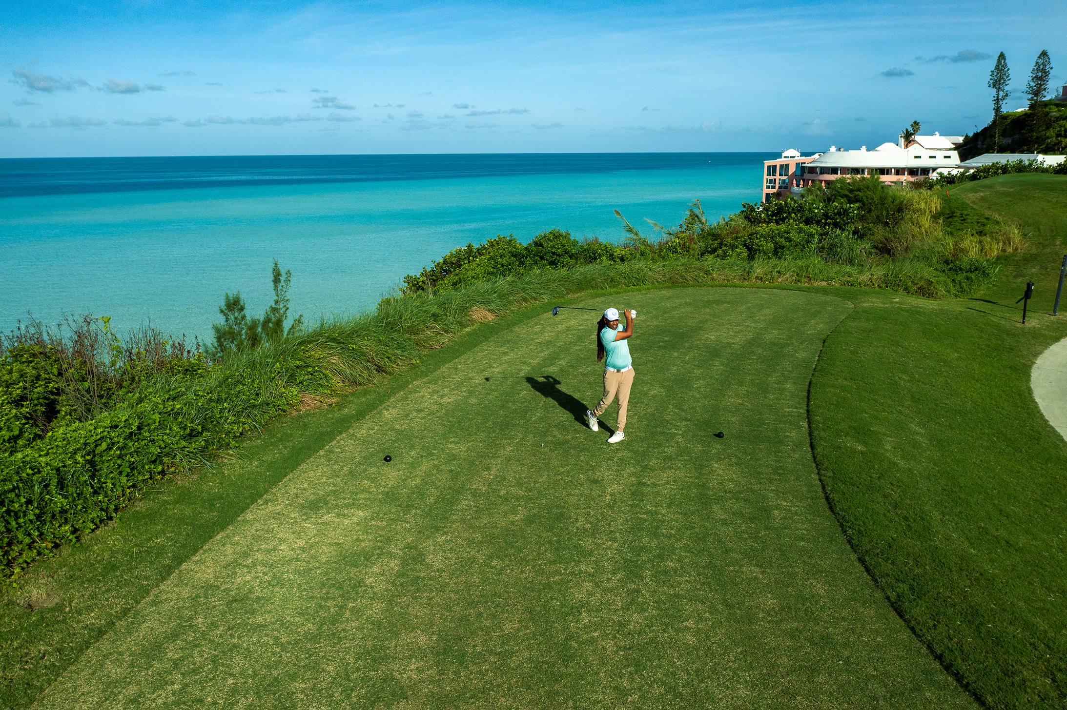 A woman playing golf on a green course by the ocean, with buildings and trees in the background.