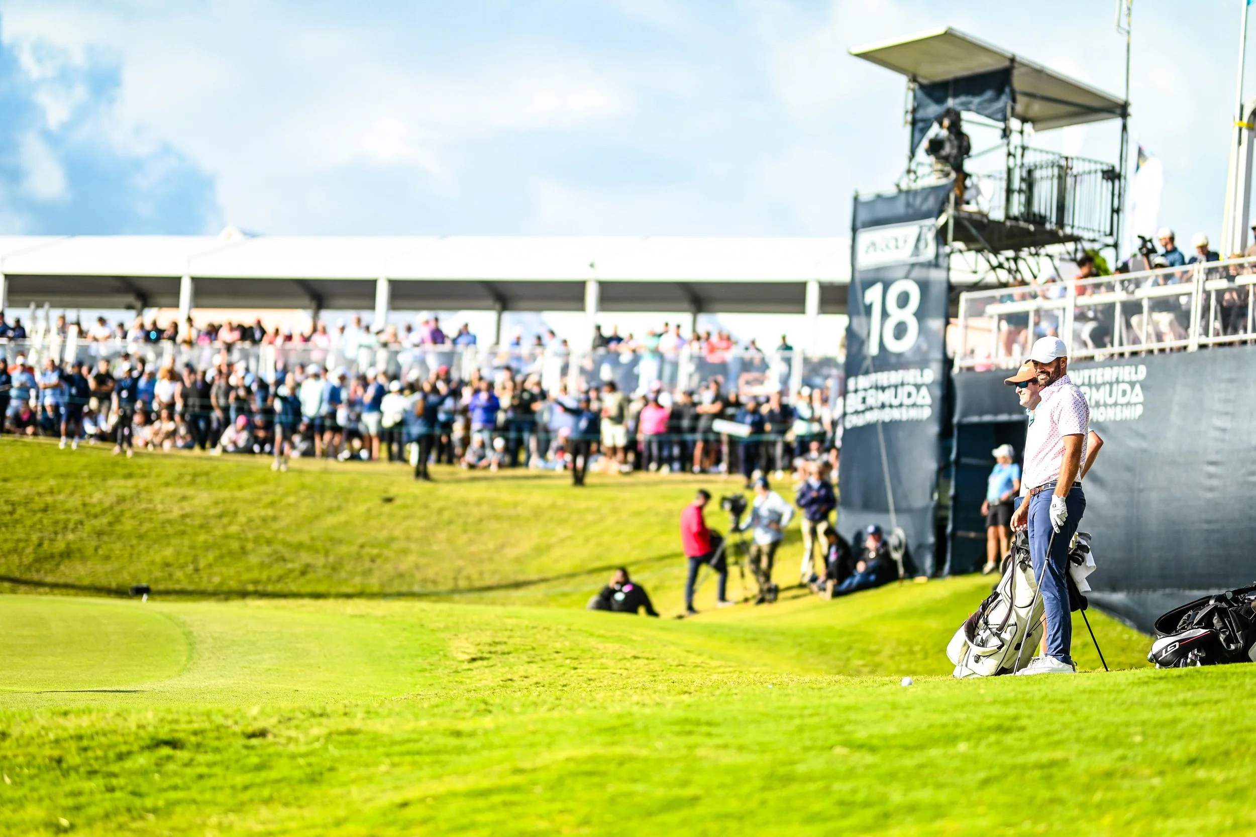 Scene of a crowded golf tournament with spectators on a viewing deck and two men with golf bags on the green, sunny weather.