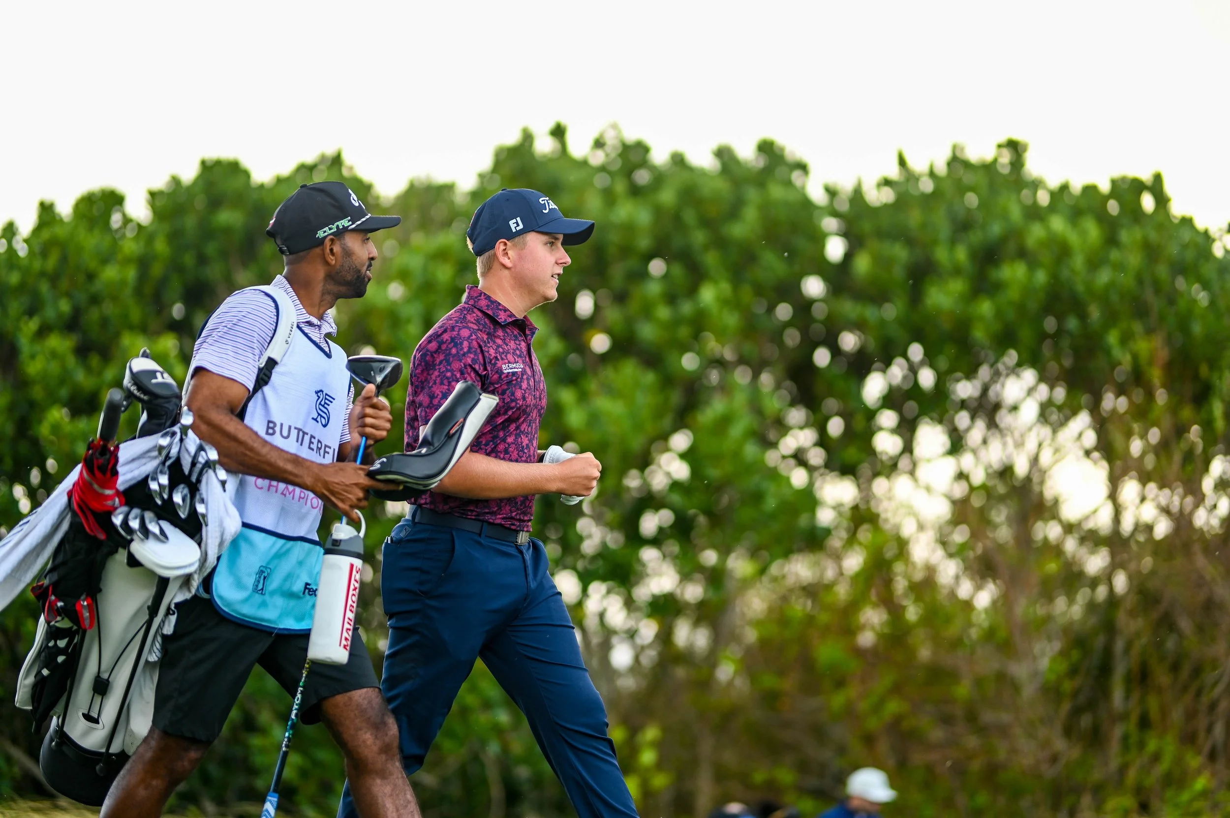 Two male golfers walking on a golf course with trees in the background, one carrying a golf bag.