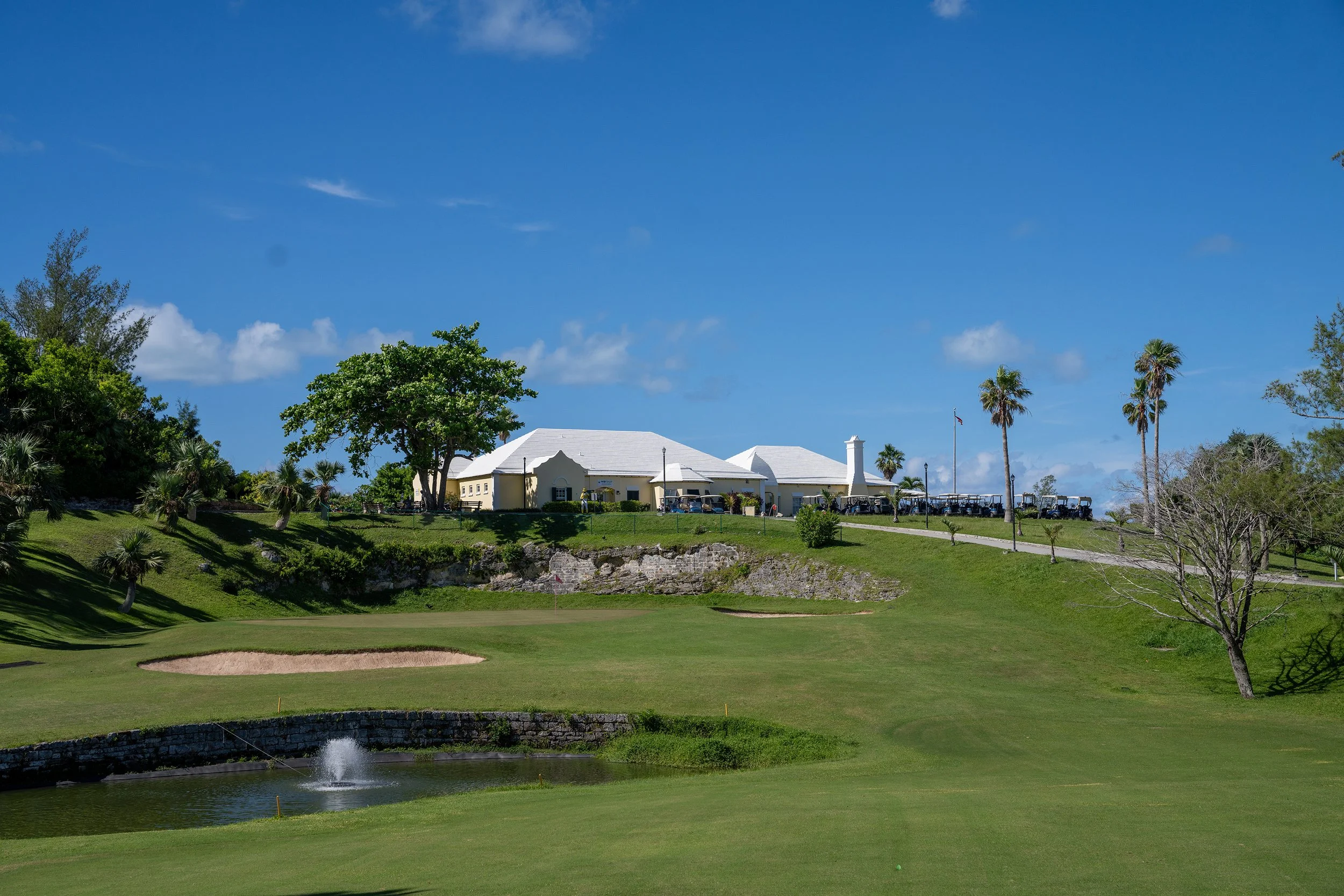 A golf course with a green, a sand trap, and a water fountain, with a white clubhouse building and palm trees in the background under a blue sky.