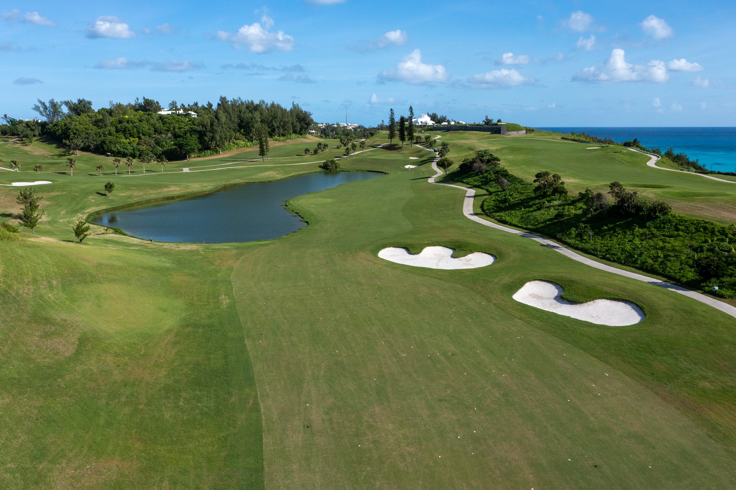 A scenic view of a golf course near the ocean, featuring green fairways, sand bunkers, a small pond, and trees with the sea in the background under a partly cloudy sky.