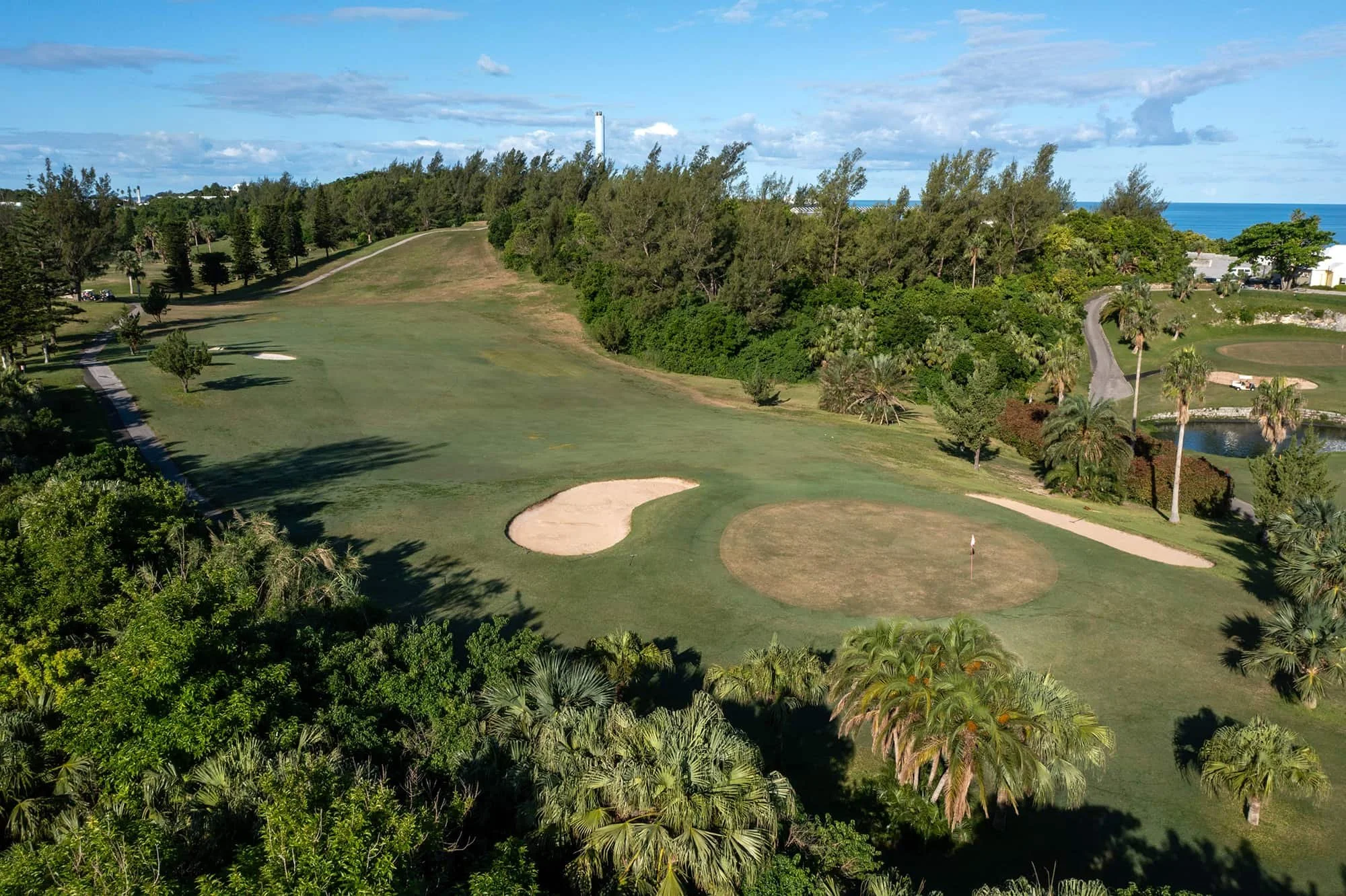 Aerial view of a golf course with sand traps, green fairways, trees, and a small pond, with the ocean visible in the background.