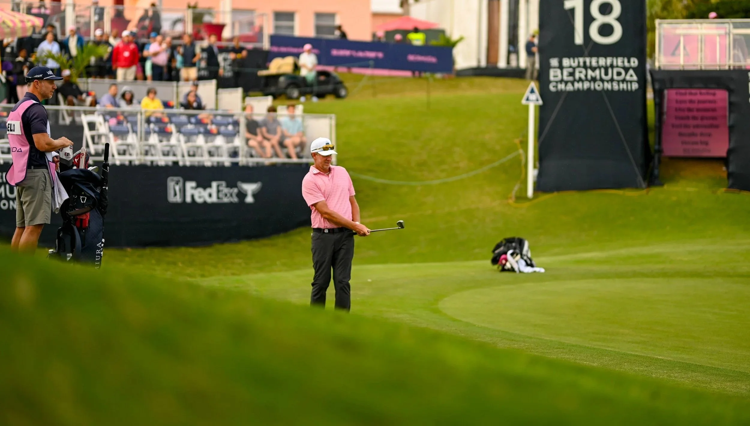 A golfer wearing a pink shirt and white cap on a golf course, preparing to take a shot, with caddies and spectators watching nearby during the Butterfield Bermuda Championship.