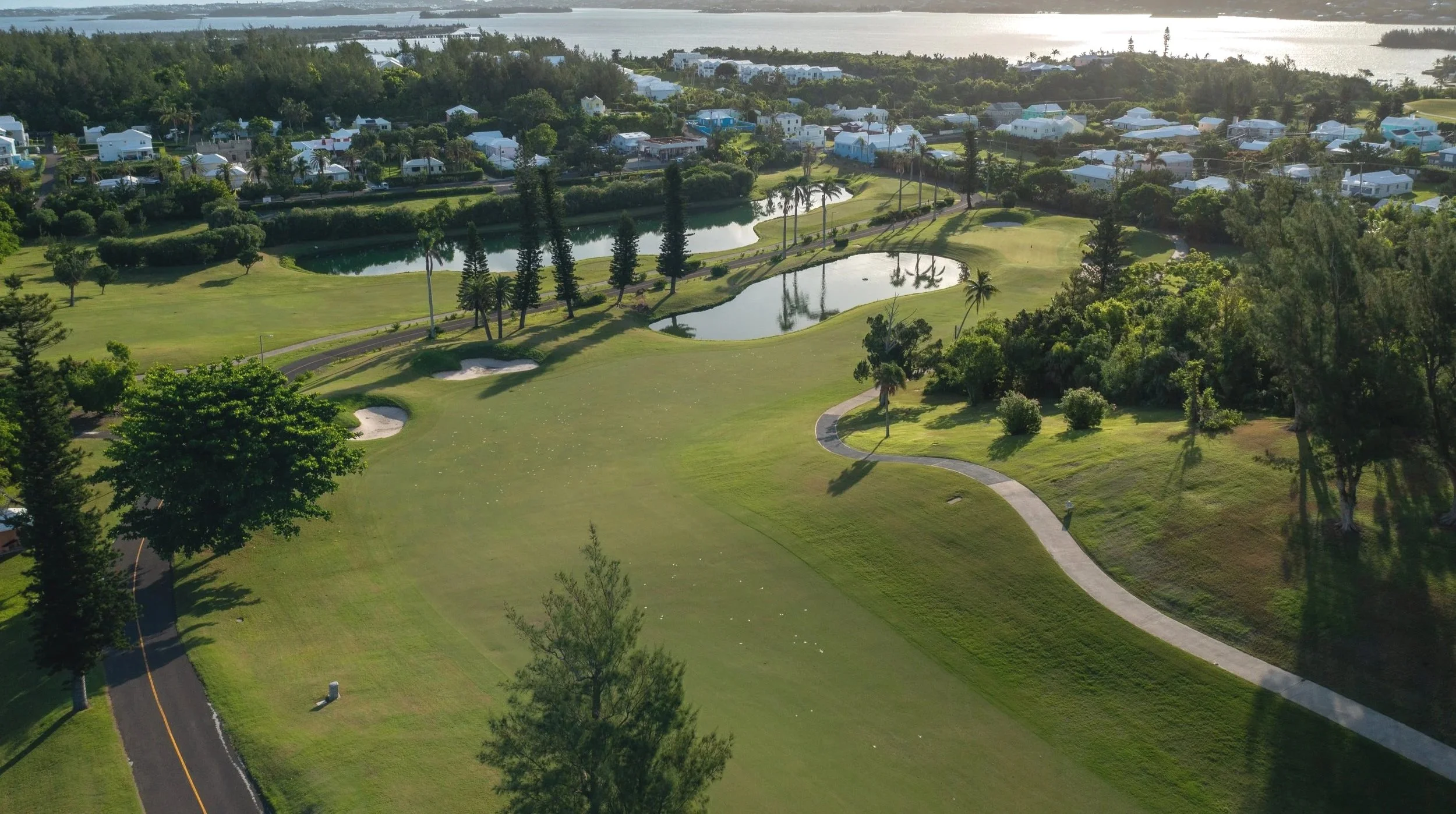 Aerial view of a golf course with ponds, trees, and a walking path, located near residential houses and water in the background.