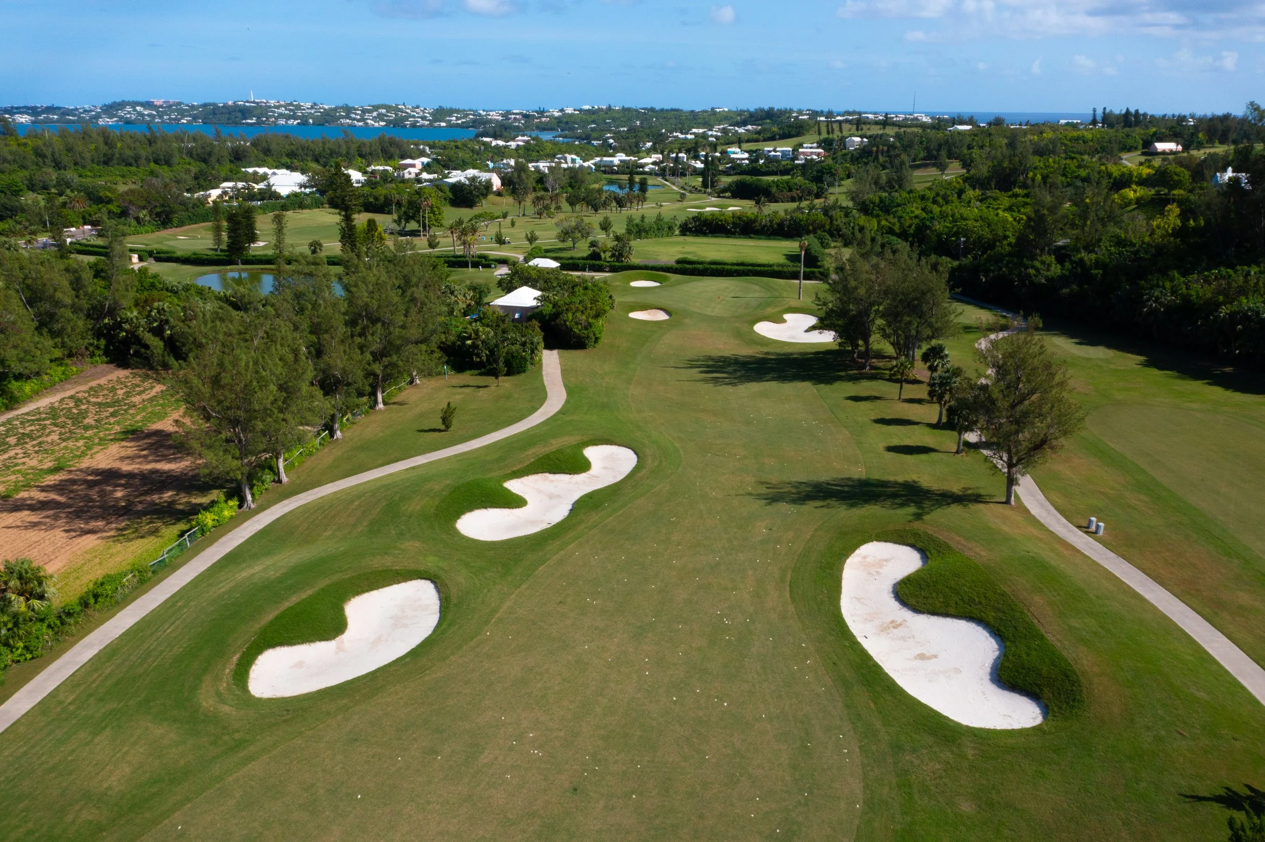 Aerial view of a golf course featuring multiple sand bunkers, green fairways, and trees, with a body of water and distant shoreline in the background.