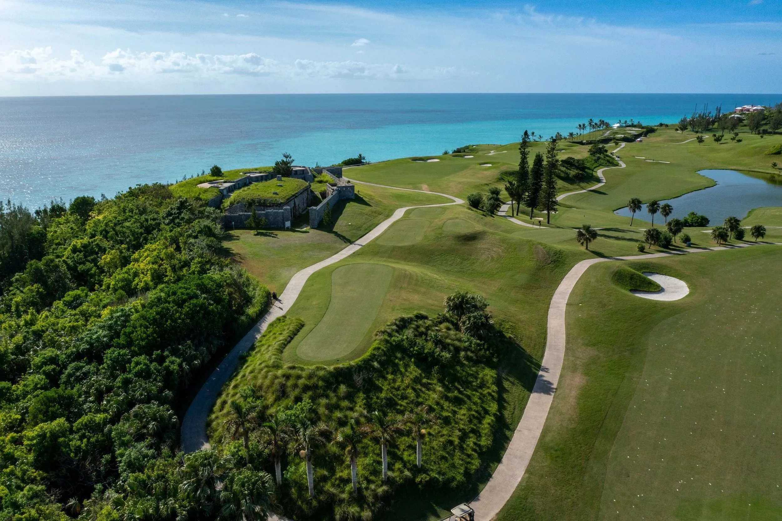 An aerial view of a golf course with green fairways, sand traps, and water hazards, near the ocean with blue sky and scattered clouds.