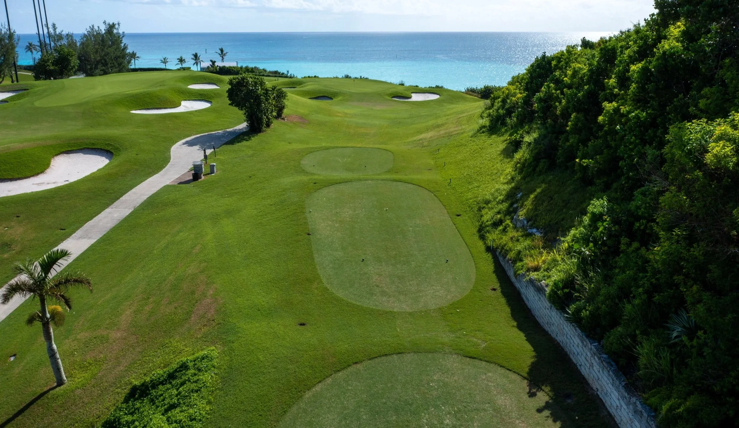 A golf course overlooking the ocean, with green fairways, sand bunkers, and tropical trees.