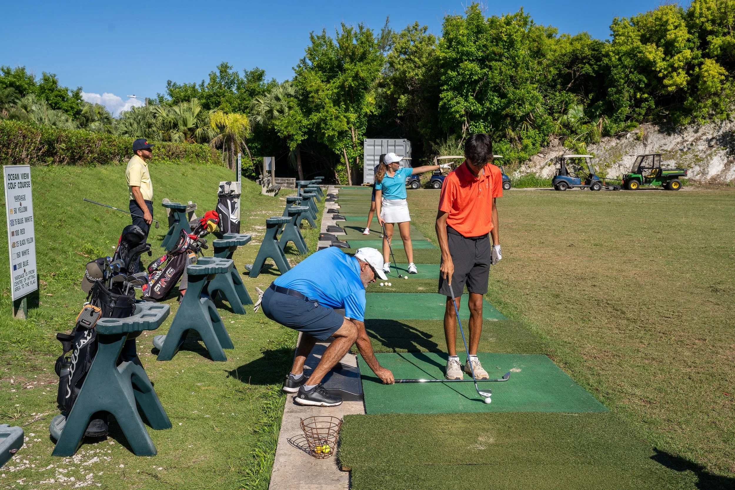 People practicing golf at an outdoor driving range with golf clubs, golf carts, and lush green trees in the background.