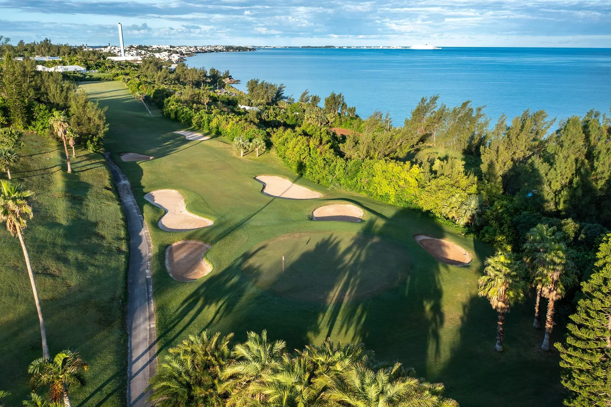 Aerial view of a golf course near the ocean, featuring sand bunkers, green fairways, numerous tall palm trees, and a distant shoreline with buildings and a cruise ship on the water.