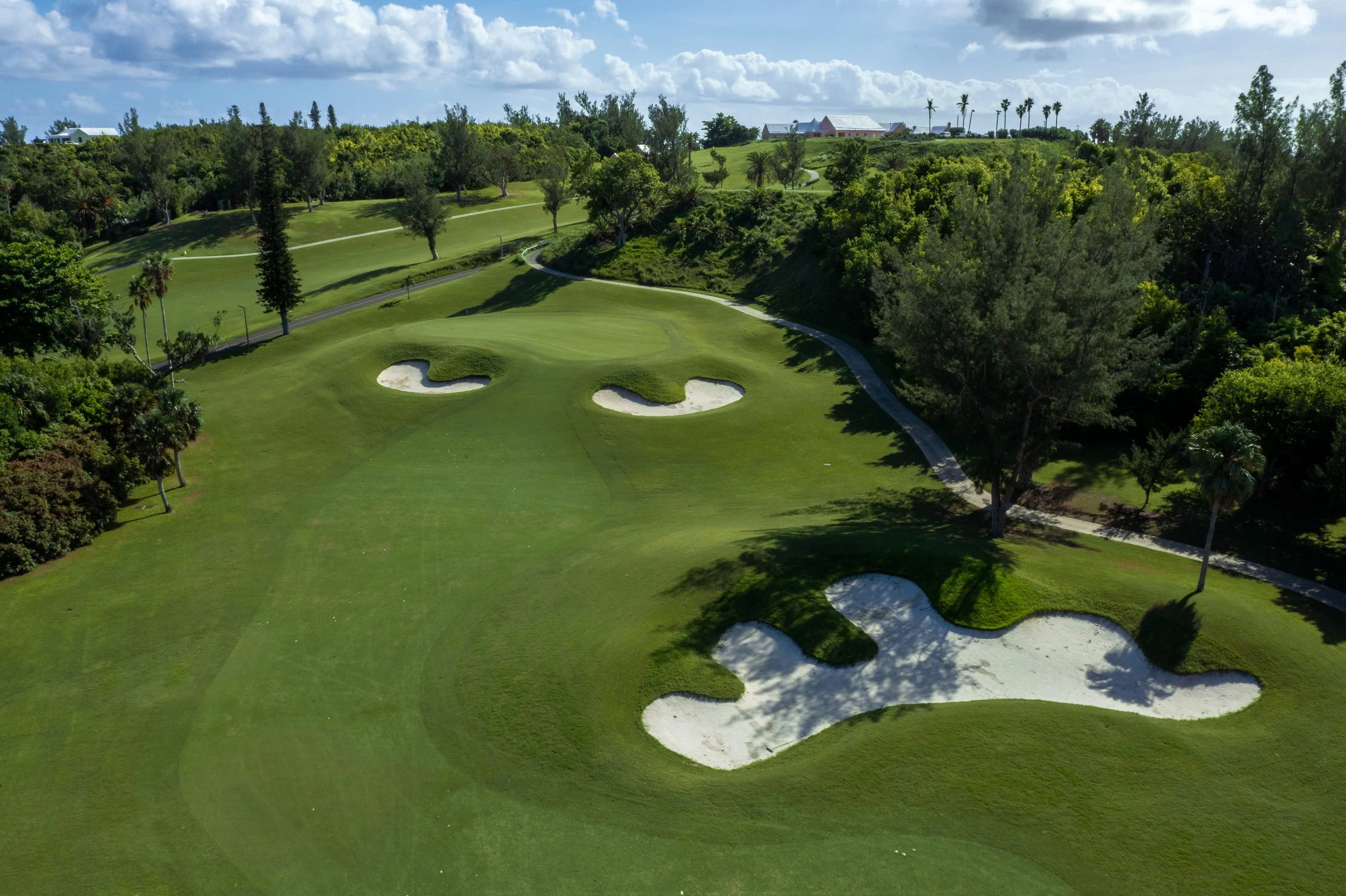 Aerial view of a golf course with green fairways, sand bunkers, and trees, with buildings and palm trees in the background under a partly cloudy sky.