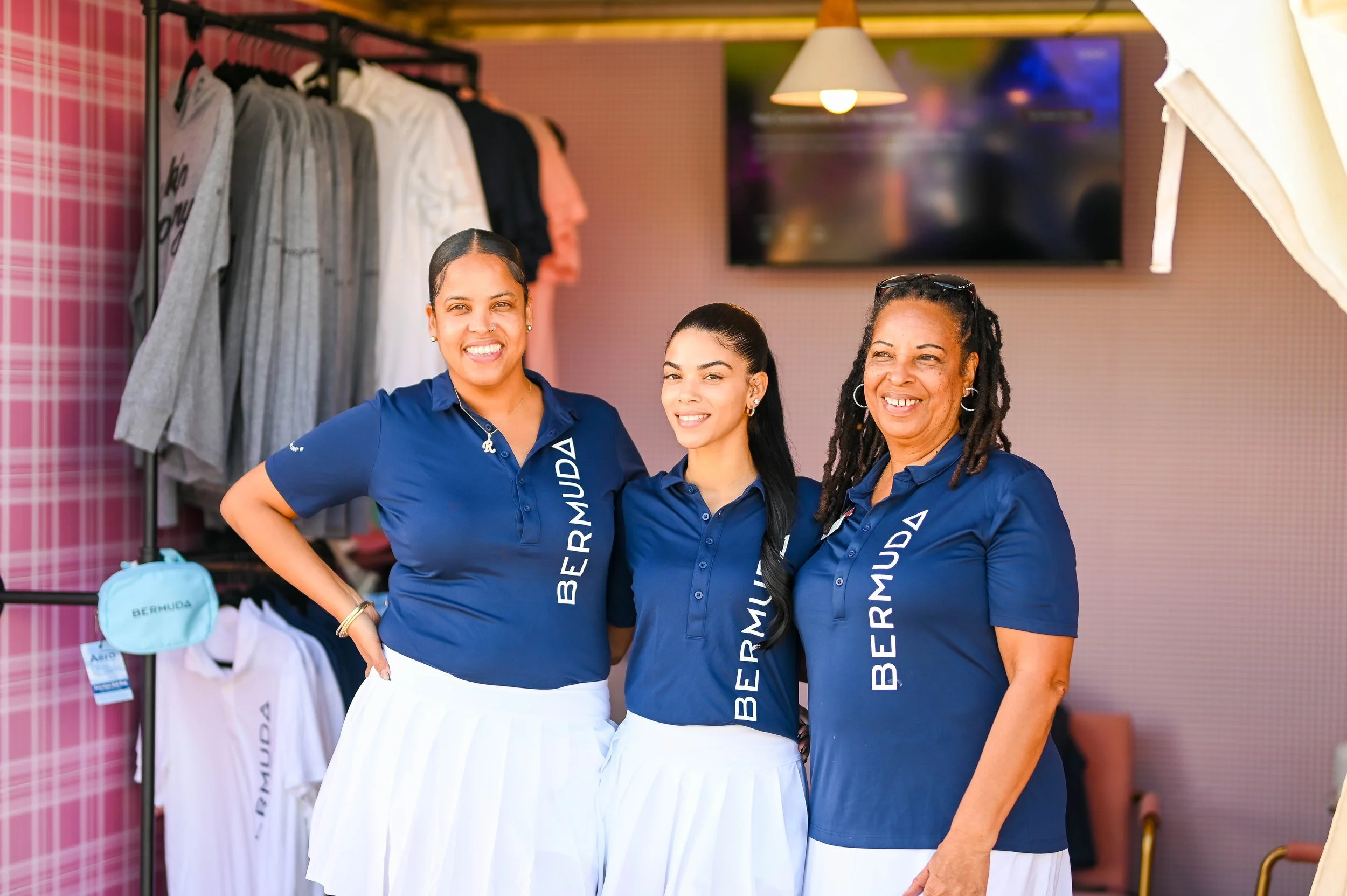 Three women wearing matching blue shirts with white skirts, smiling, standing together in a retail store with clothing racks in the background.