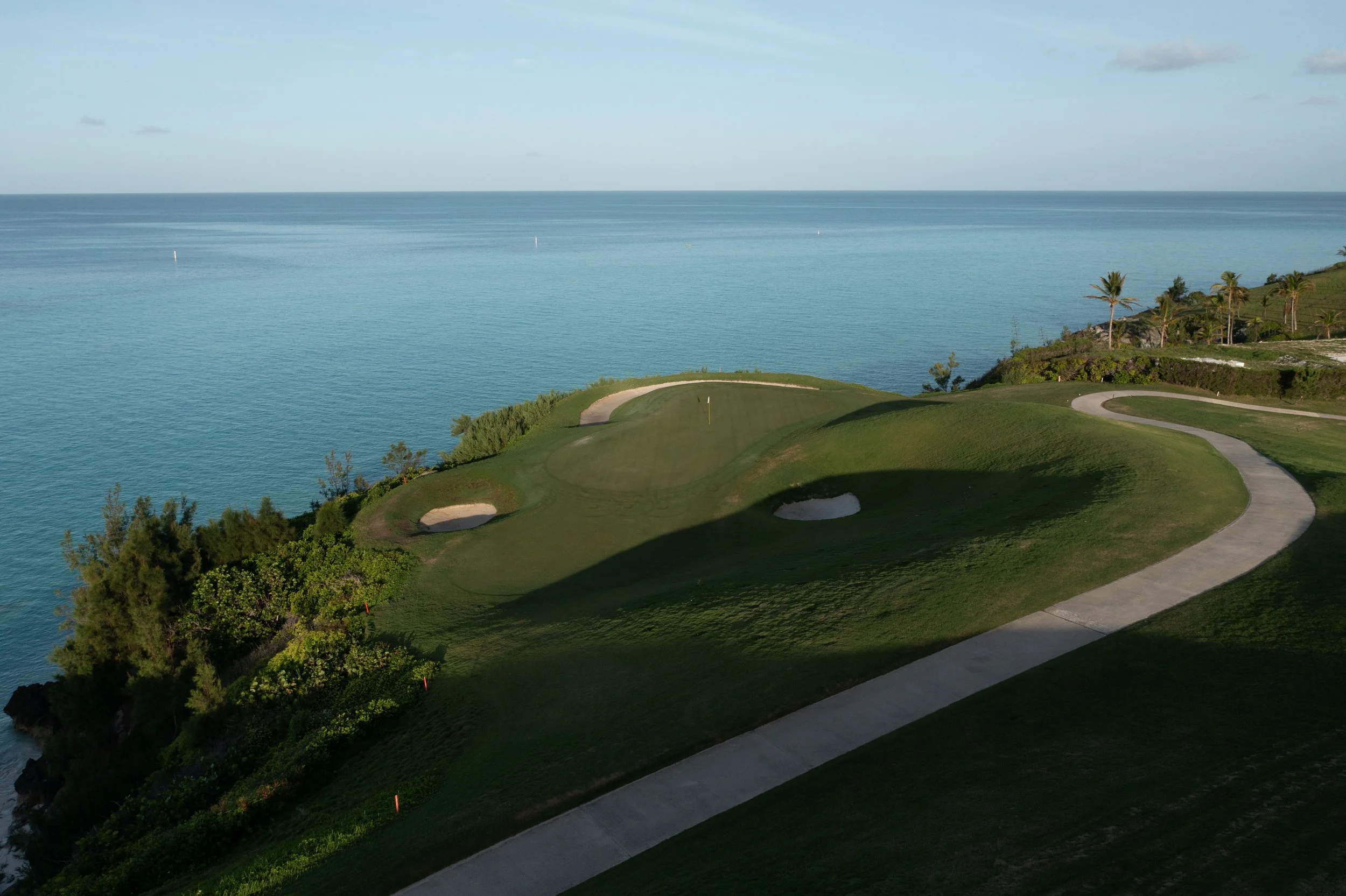 A scenic view of a golf course located near the ocean, with green fairways, sand bunkers, a winding pathway, and palm trees, under a partly cloudy sky.