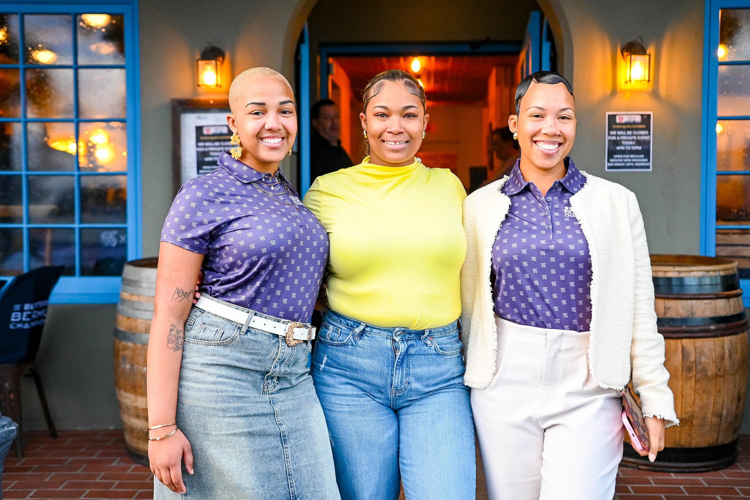 Three women standing close together smiling outside a restaurant or cafe with warm lighting, barrels, and windows behind them.
