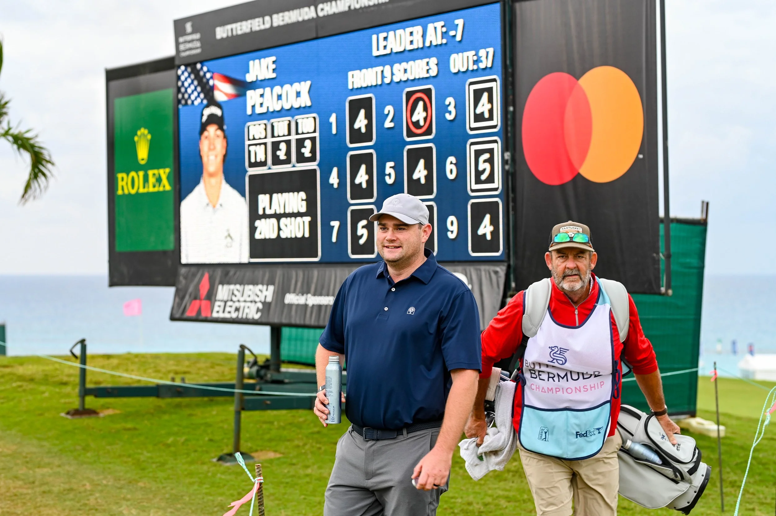 Two men walking on a golf course near a large scoreboard displaying scores from a Bermuda Championship golf tournament. One man is dressed in a navy shirt and gray pants, holding a water bottle, and the other is wearing a white and red golf outfit with a hat and sunglasses, carrying a golf bag.