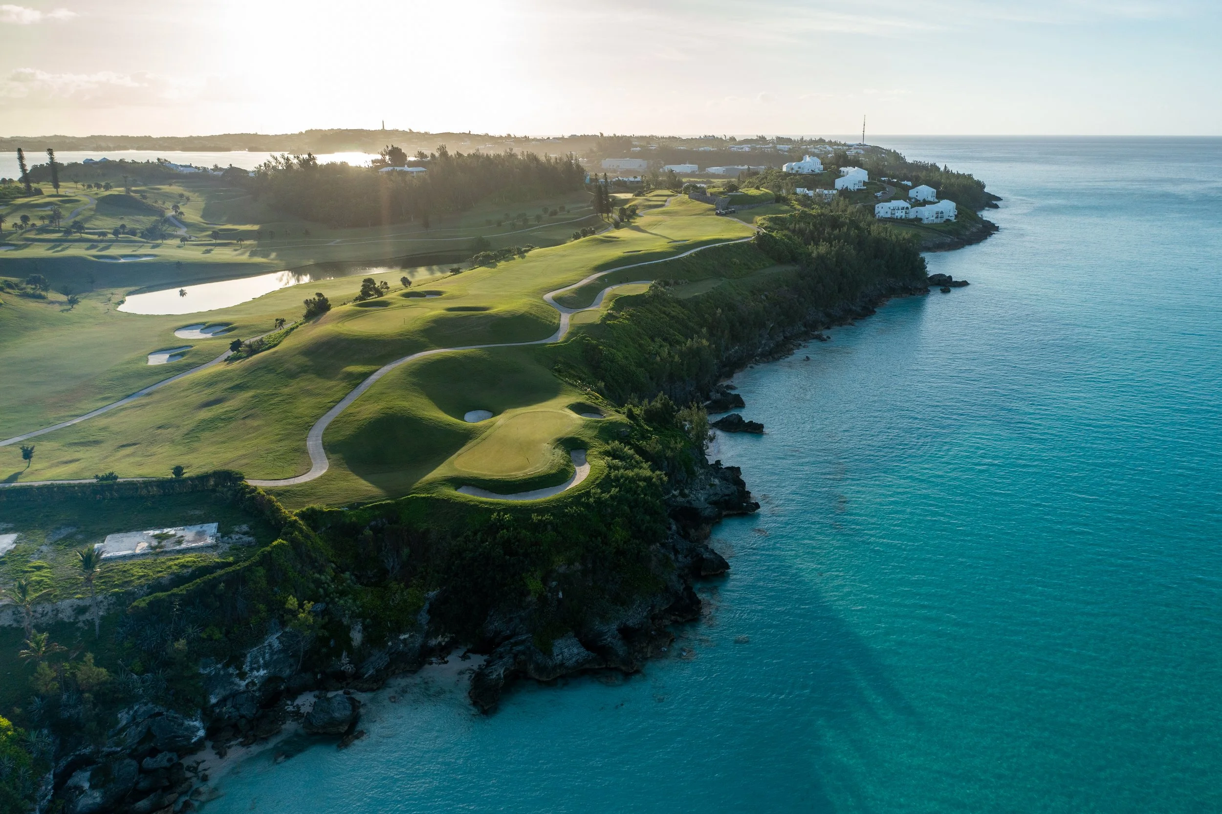 Aerial view of a coastal golf course with green fairways and sand traps, overlooking the ocean with rocky shorelines and white buildings along the coast, under a partly cloudy sky.