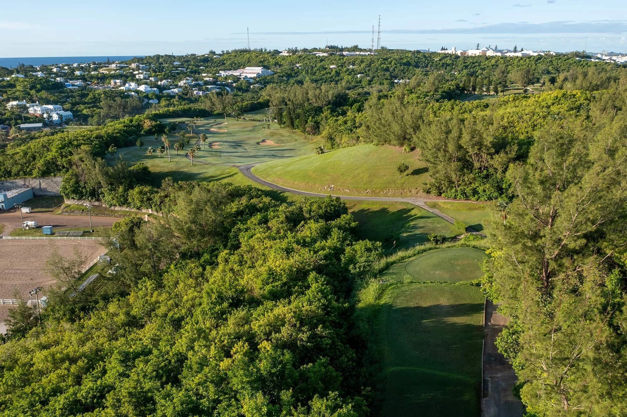Aerial view of a golf course surrounded by lush green trees and residential area in the background.