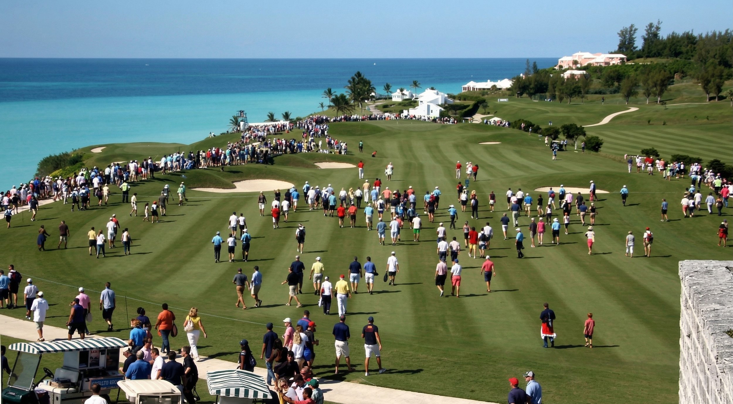 Crowds of people walking and standing on a golf course near the coast, with the ocean visible in the background. Some people are near sand traps and golf carts are in the foreground.